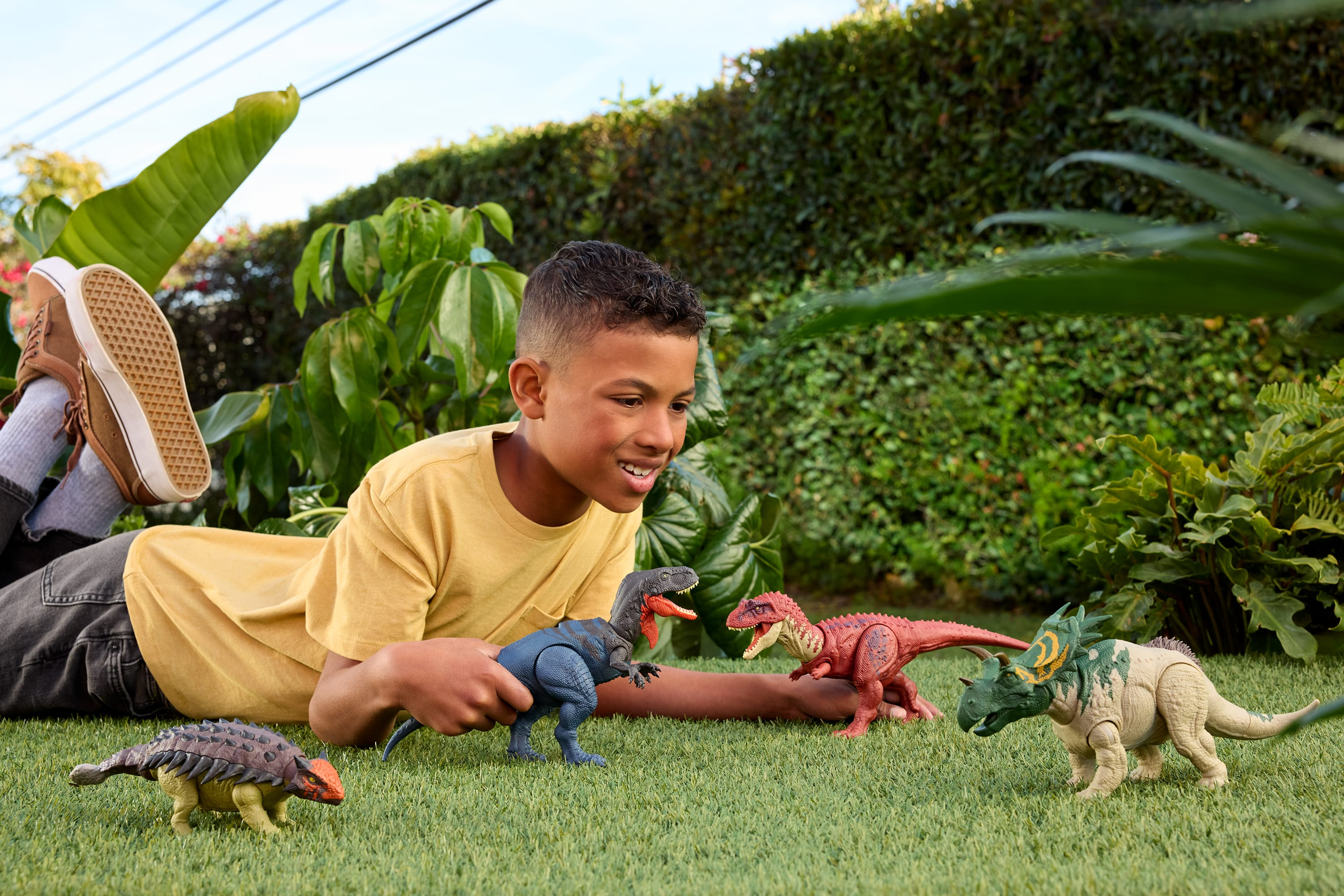 Chłopiec leży na trawie, bawiąc się zabawkami dinozaurów. Tło obejmuje zielone liście i buty.