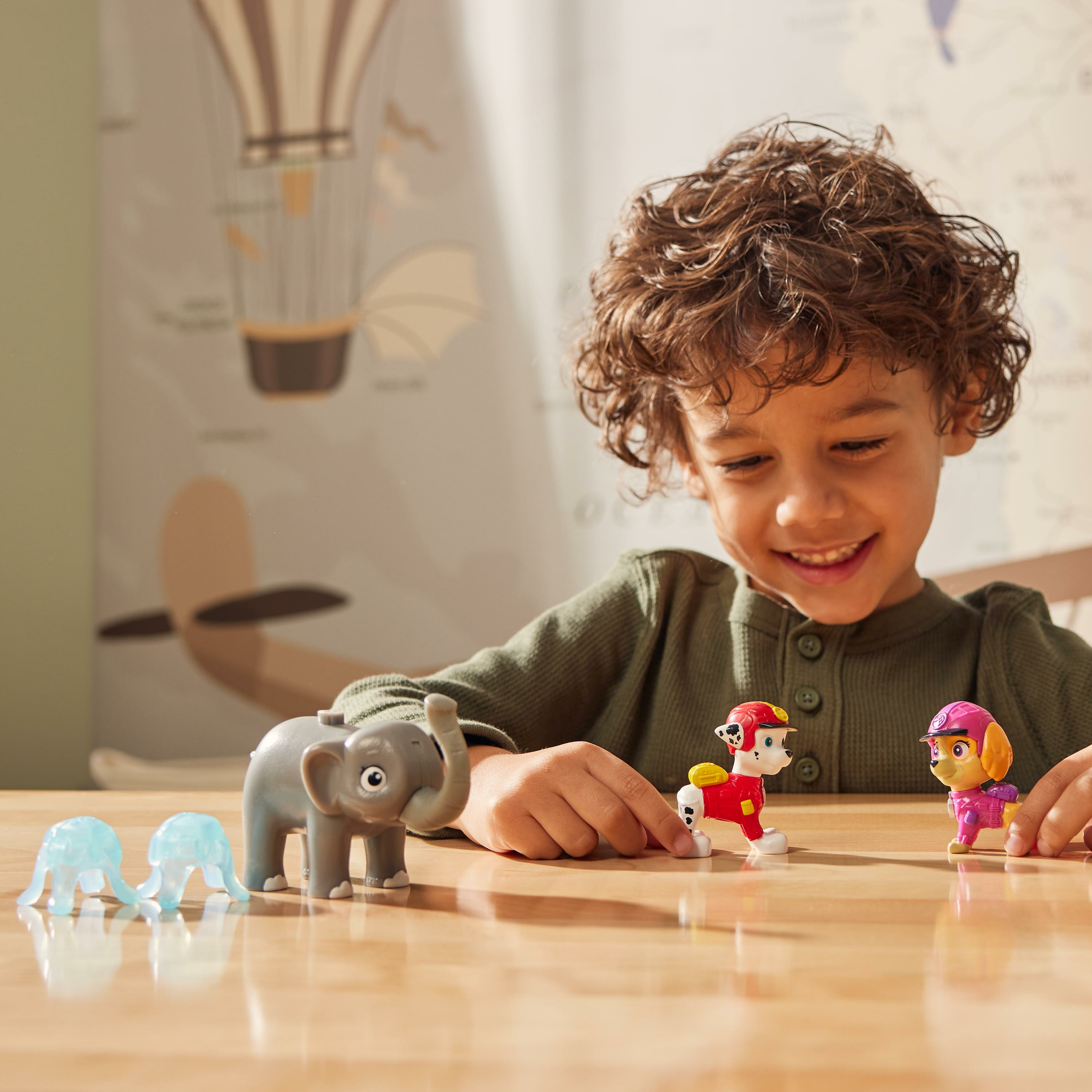 Un bambino sorride, gioca con dei giocattoli su un tavolo. I giocattoli includono un elefante grigio, personaggi di PAW Patrol e meduse blu.