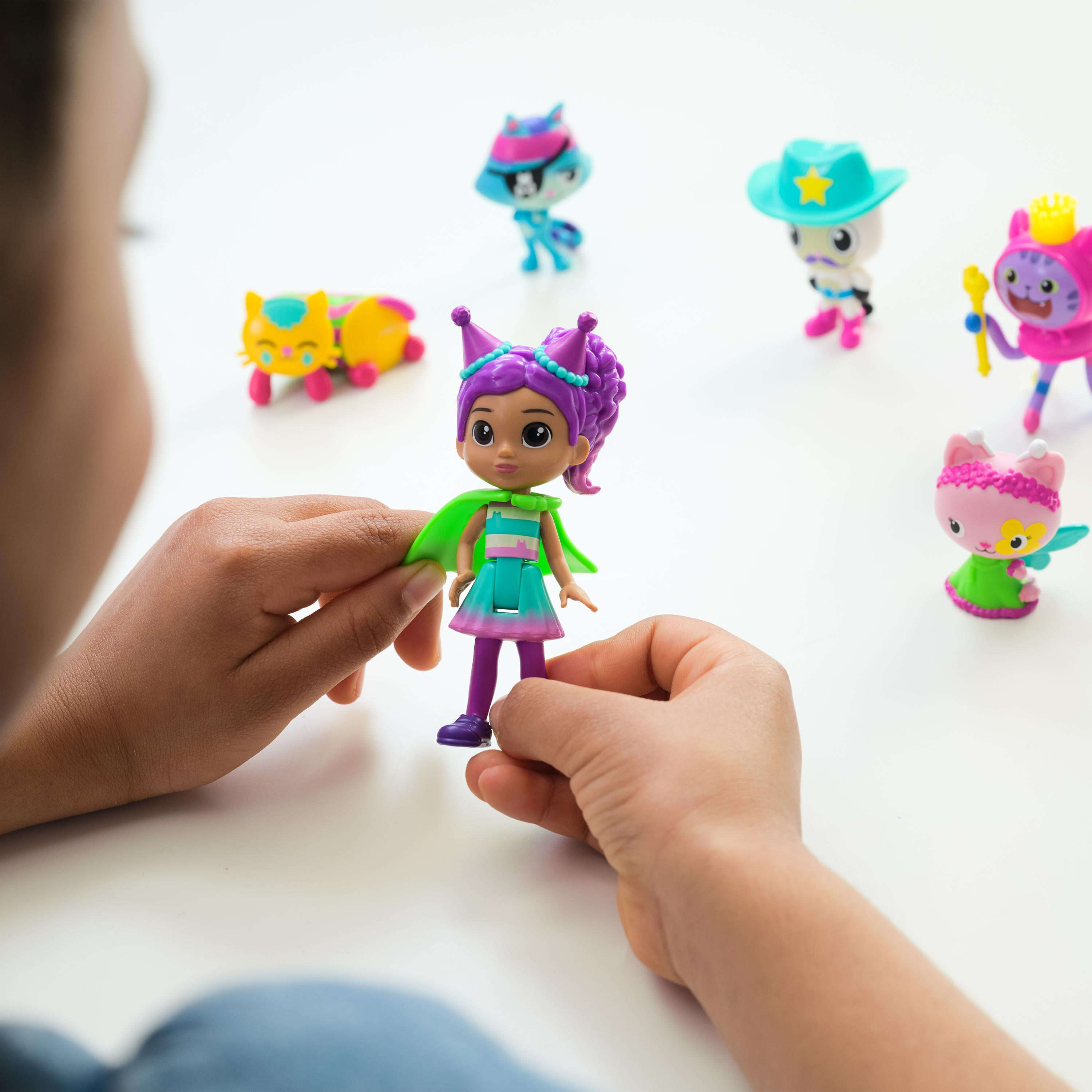 Osoba trzyma lalkę z fioletowymi włosami i zielonym płaszczem. Inne lalki w kształcie kota są w tle.