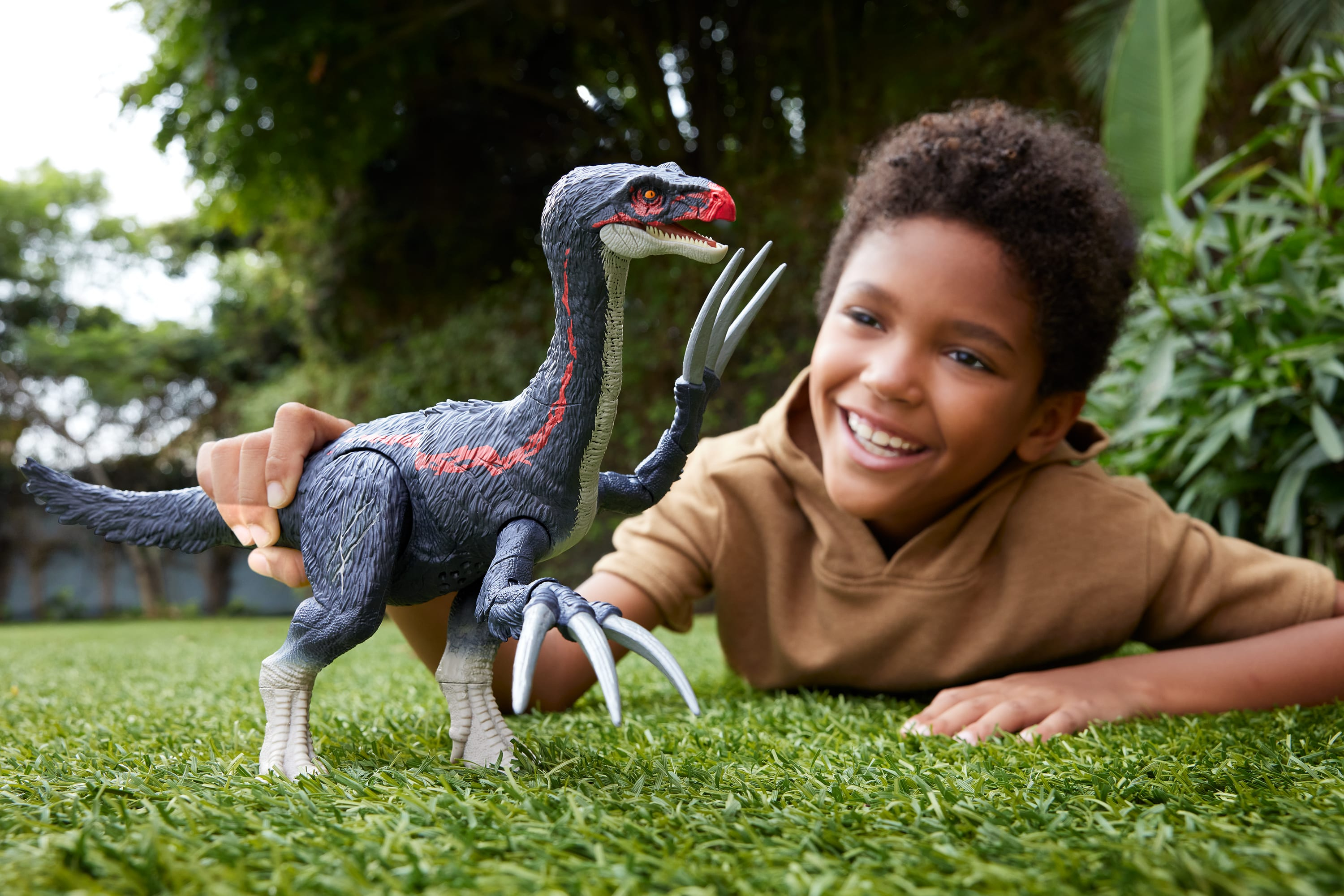 A boy smiles, holding a toy dinosaur on grass. Green trees in the background. The dinosaur is gray and red.
