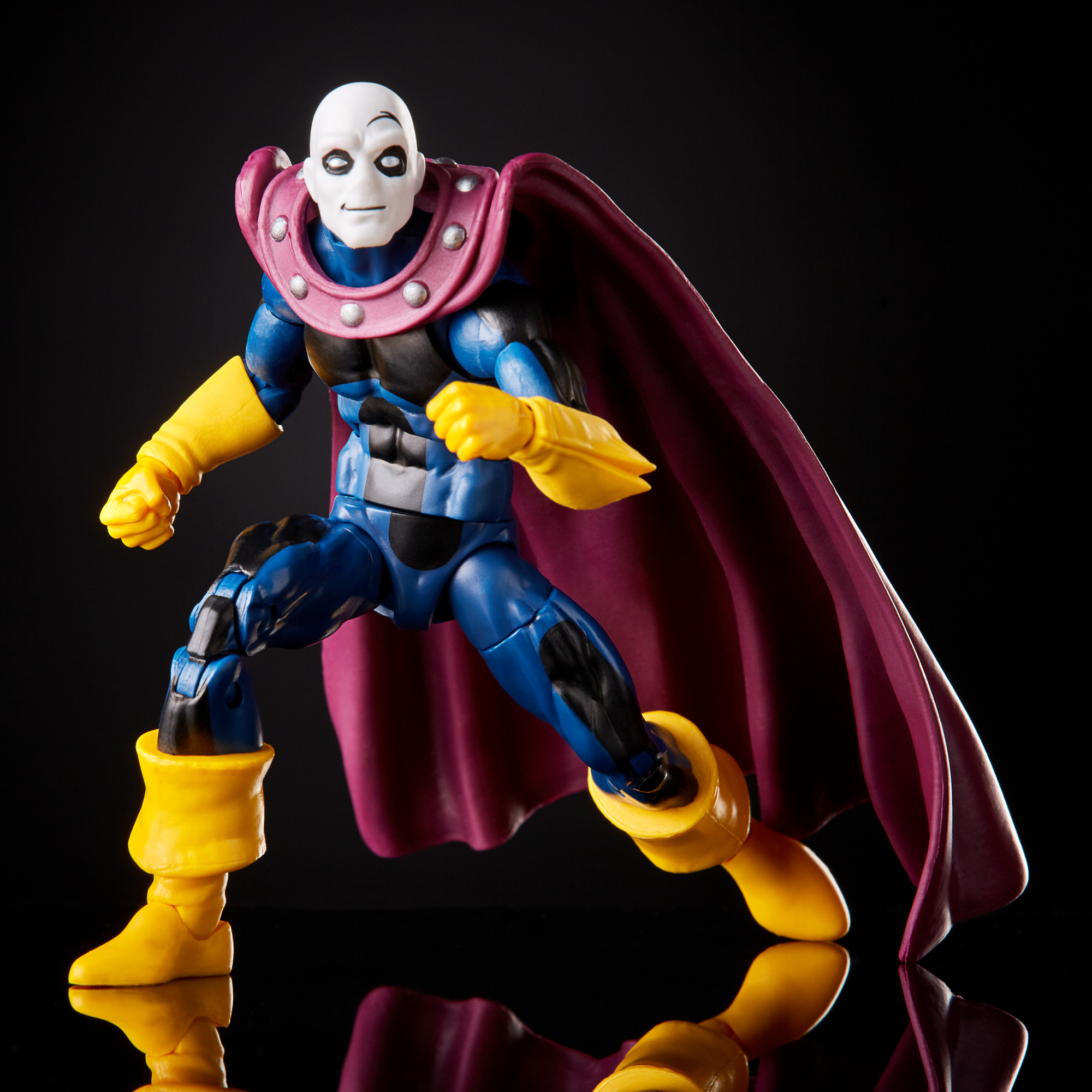 Action figure di un personaggio con viso bianco, tuta blu, guanti e stivali gialli e mantello viola, su sfondo nero.