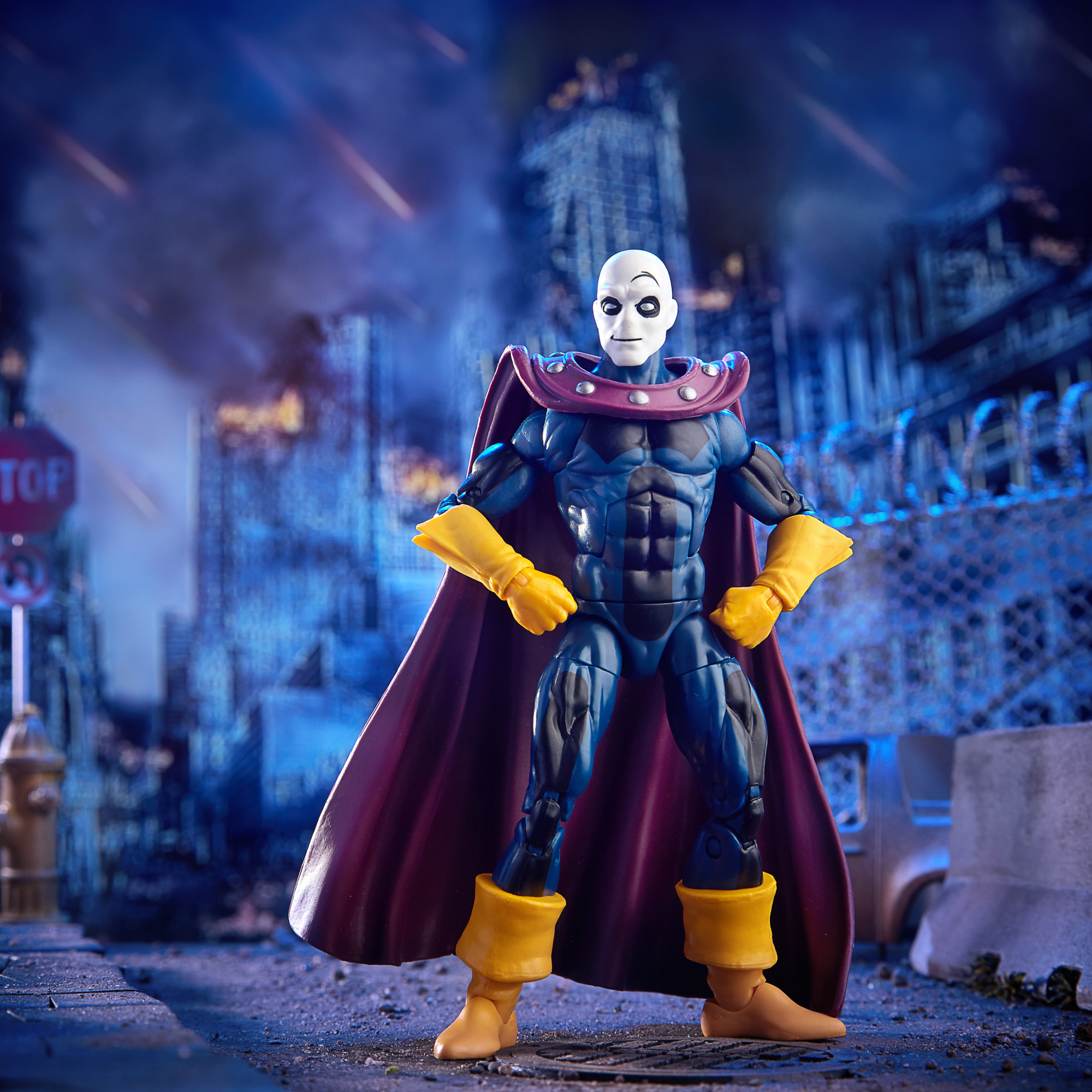 Action figure di un uomo calvo in abito e mantello blu, guanti e stivali gialli, in piedi in una città distrutta.