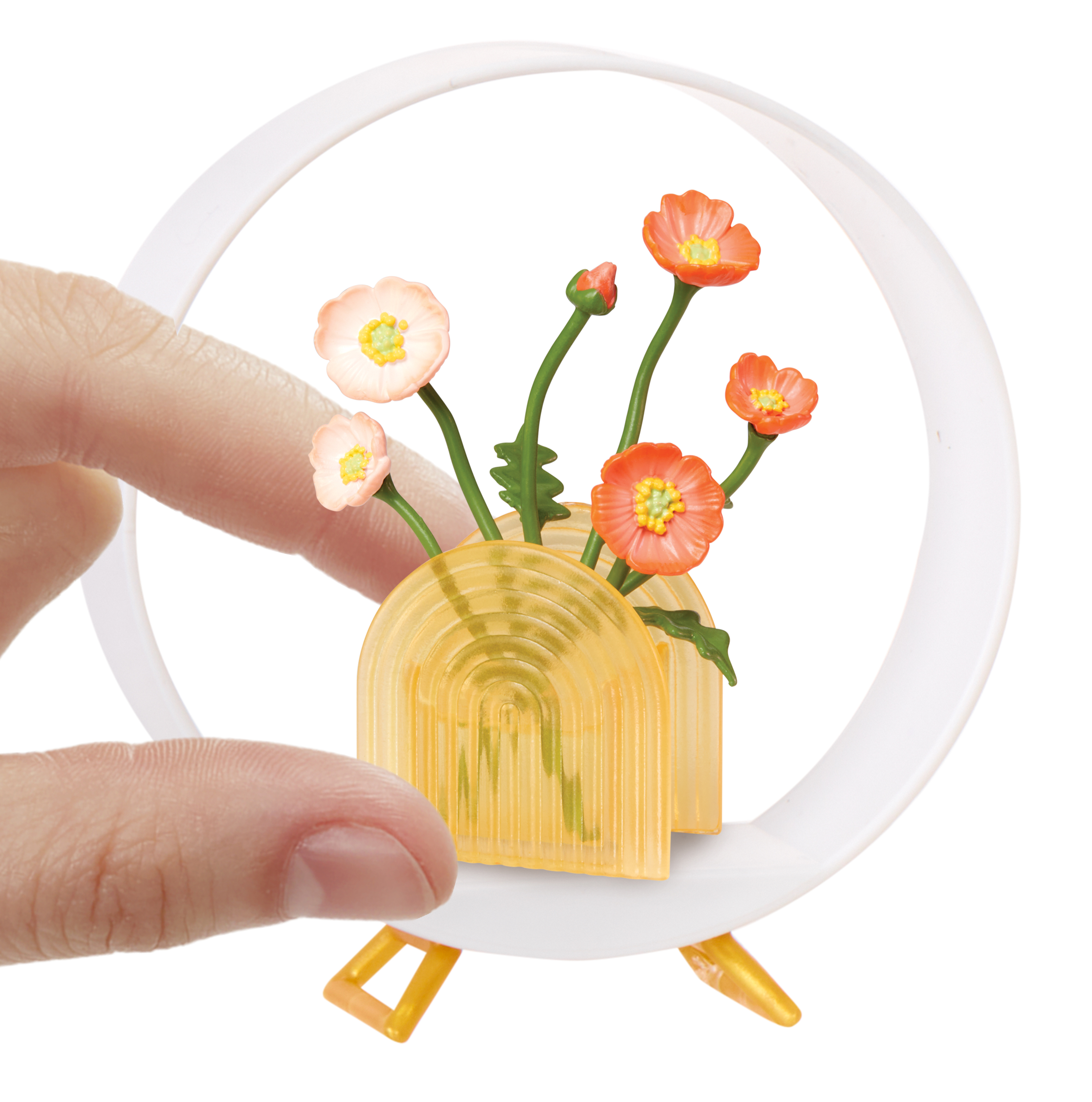 Ręka trzyma miniaturową scenę z kwiatami w żółtym wazonie, biały pierścień otacza scenę.