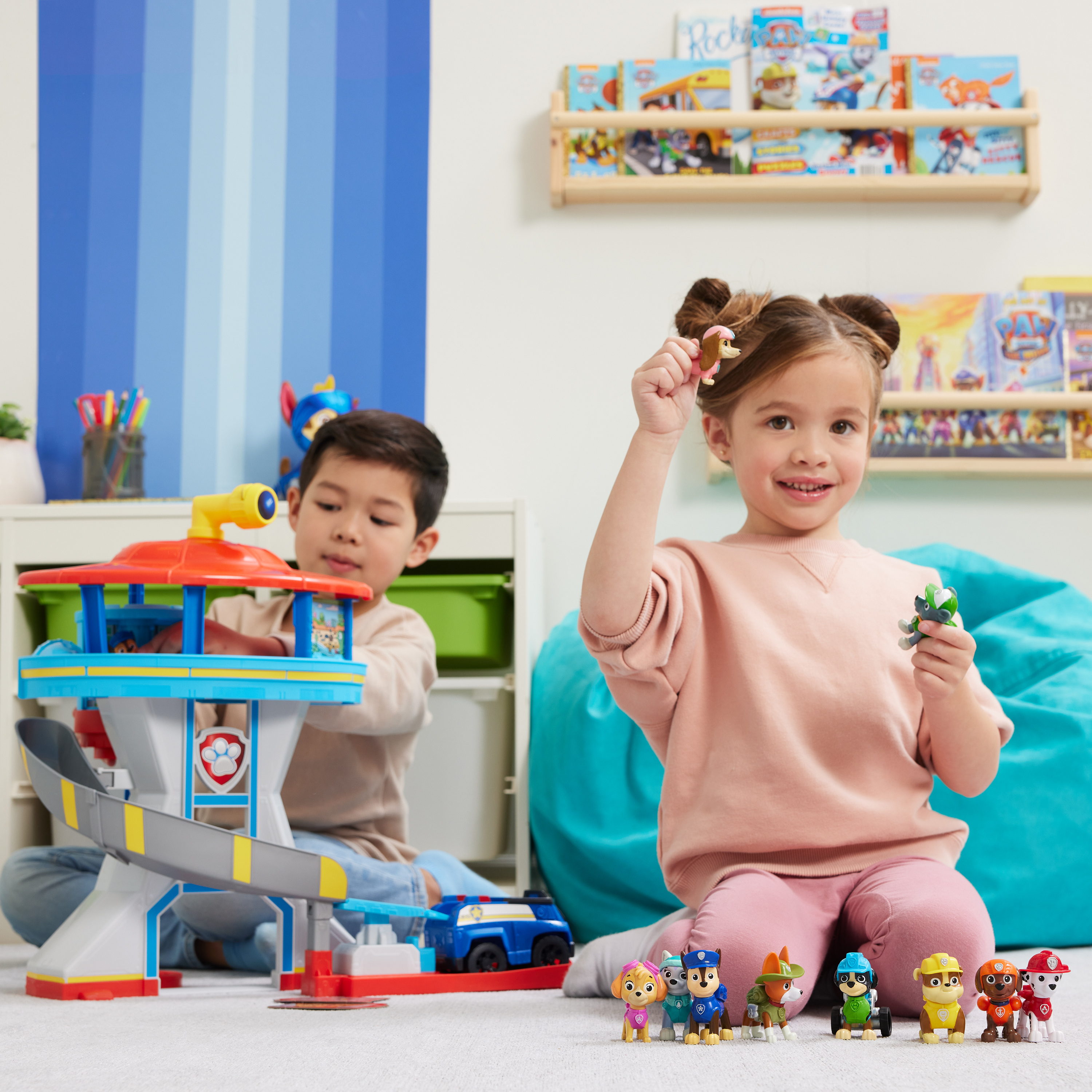 Dwoje dzieci bawiących się zabawkami z Psiego Patrolu. Jedno trzyma figurkę. Drugie bawi się wieżą i zabawką samochodem.