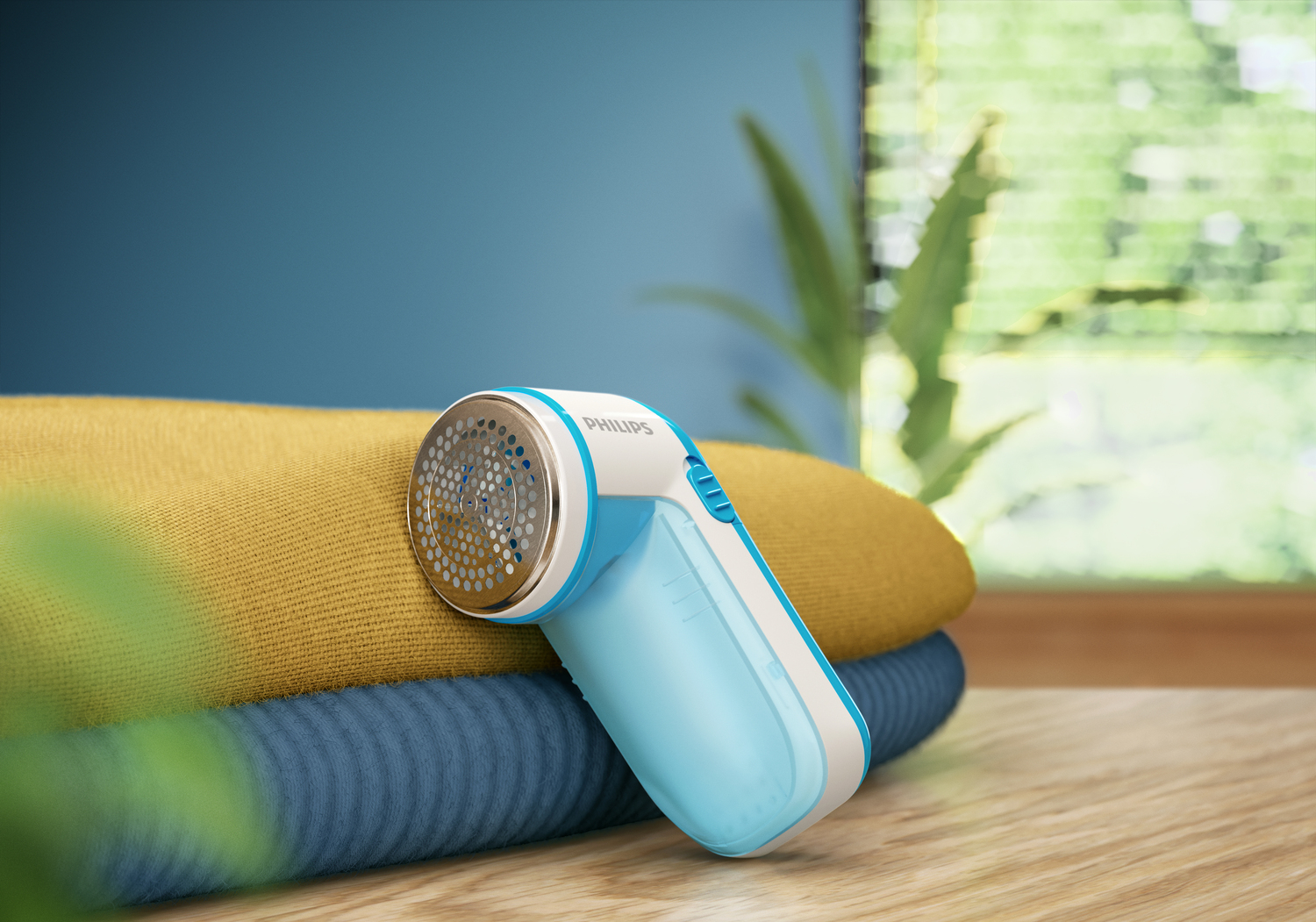 Una afeitadora de tela Philips con detalles blancos y azules descansa sobre telas amarillas y azules dobladas. Una ventana está en el fondo borroso.