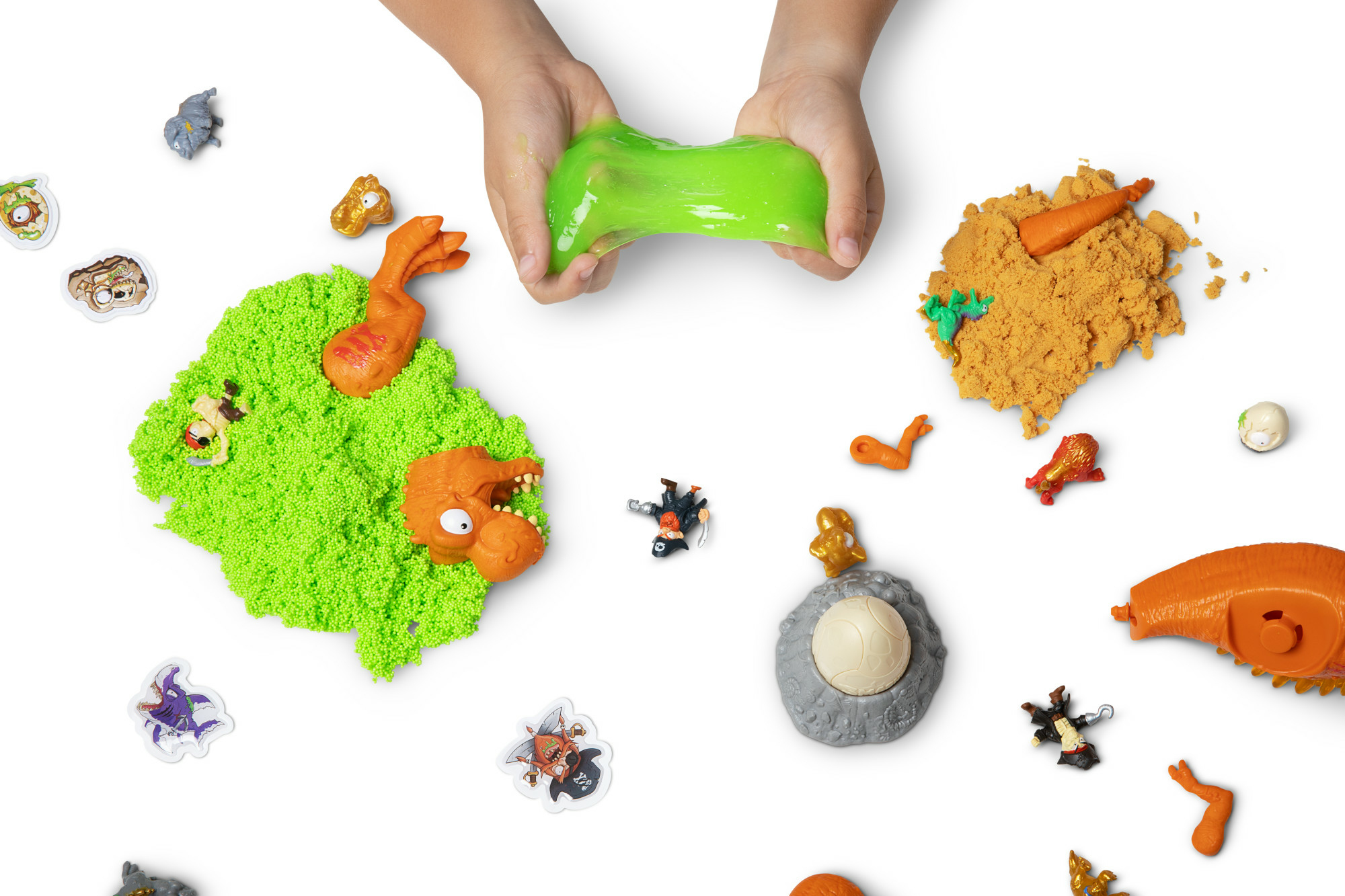 Hände halten grünen Schleim. Spielzeug und Spielsand sind auf einer weißen Oberfläche verstreut. Das Bild ist von oben.