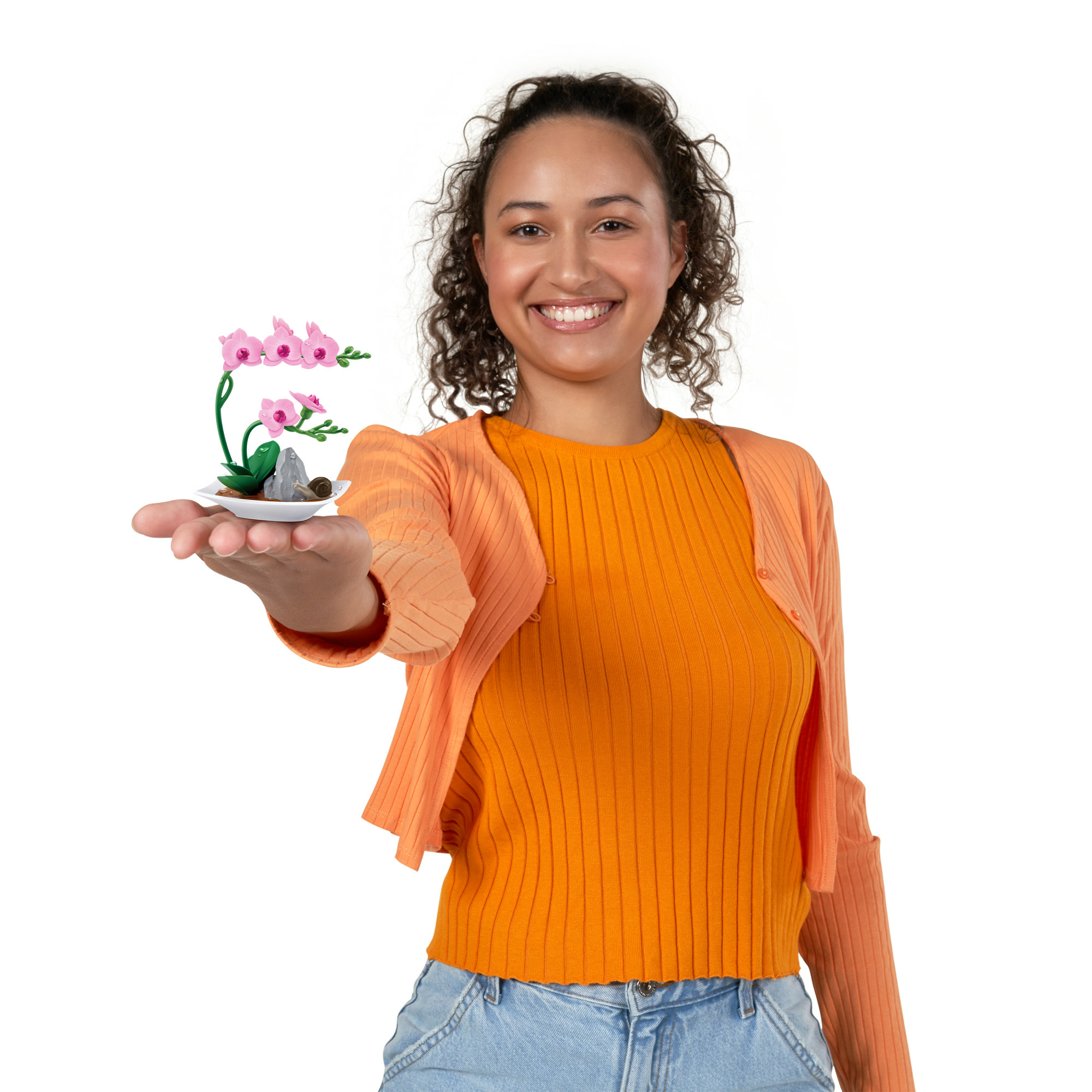 Mujer sosteniendo una orquídea en miniatura. Lleva top y cárdigan naranja, y vaqueros. Fondo blanco.