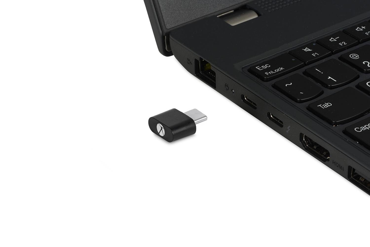 Un adaptador USB-C negro junto a un portátil. El adaptador está sobre una superficie blanca. Se ve el teclado.
