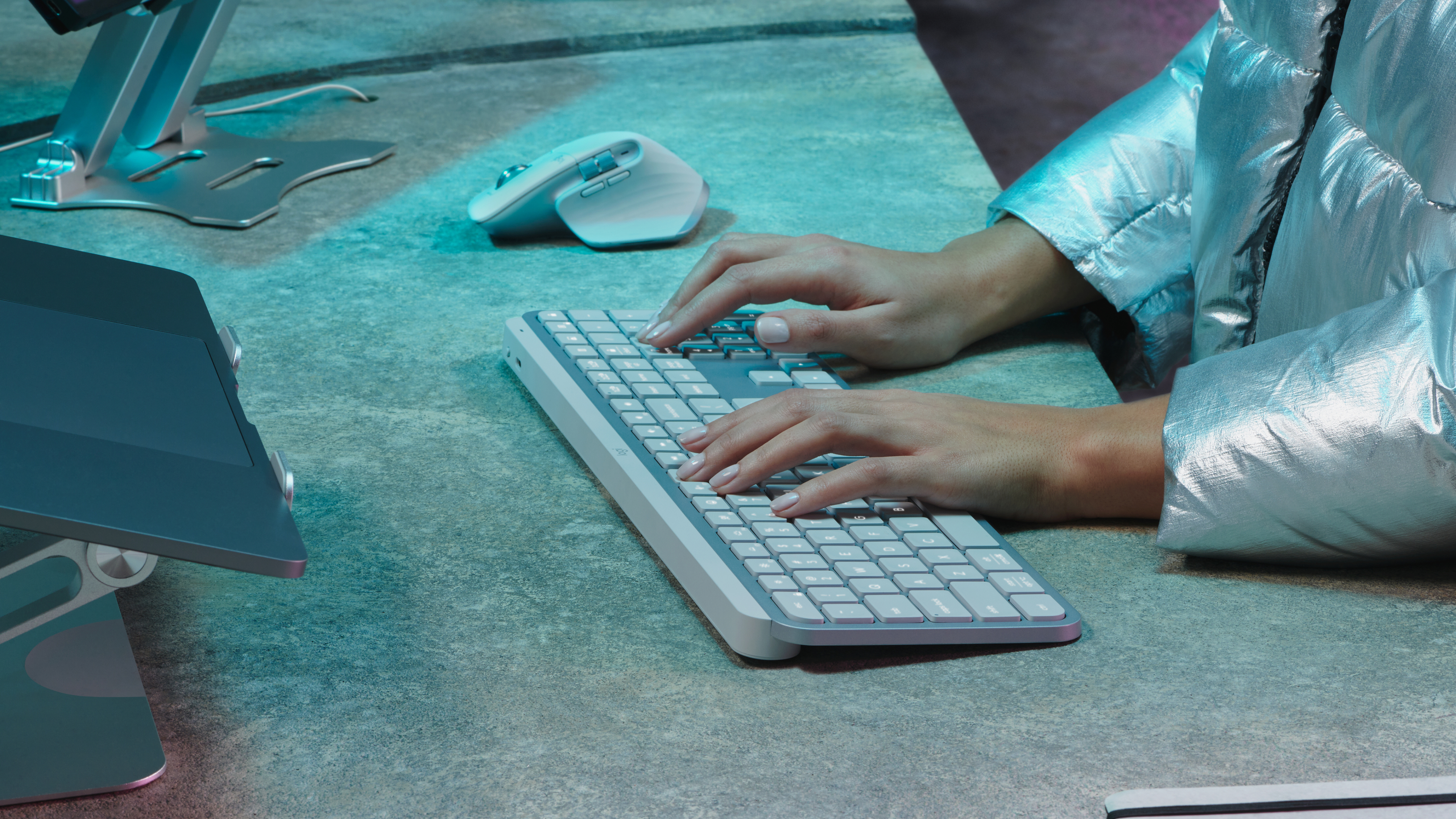 Ręce piszące na białej klawiaturze. Widoczna srebrna kurtka. Mysz, stojak na laptopa i laptop na biurku.