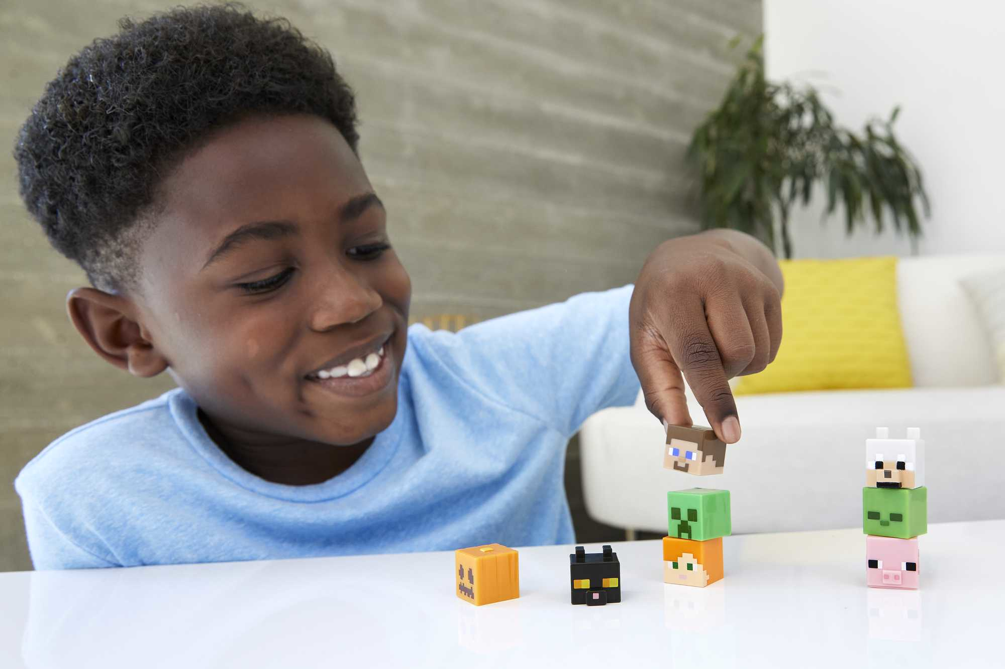 Ein lächelnder Junge stapelt Minecraft-Spielzeugblöcke. Mehrere Blöcke sind auf einem weißen Tisch angeordnet. Der Hintergrund umfasst ein Sofa und Pflanzen.