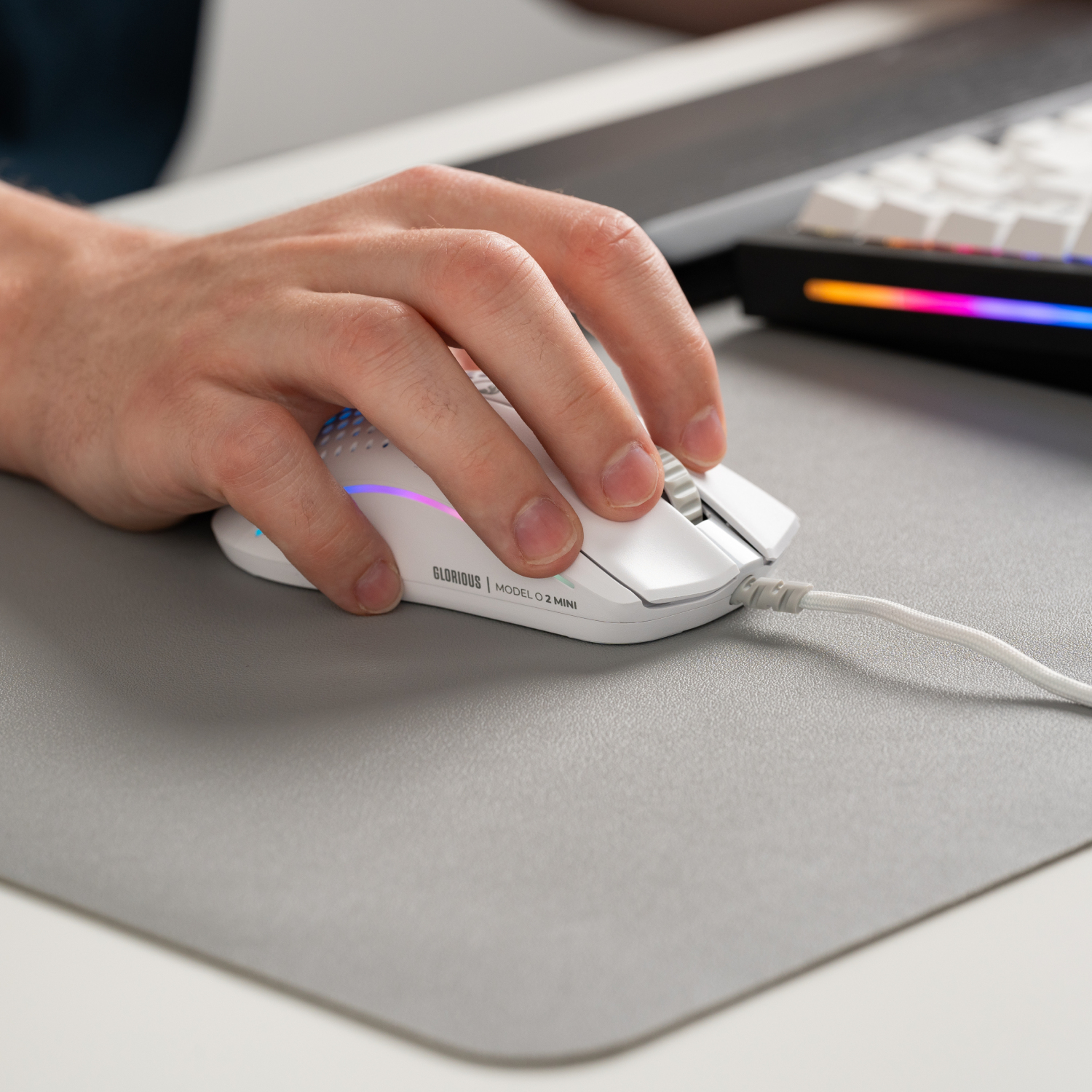 Dłoń używa białej myszy Glorious Model O 2 Mini na szarej podkładce. Klawiatura widoczna w tle.