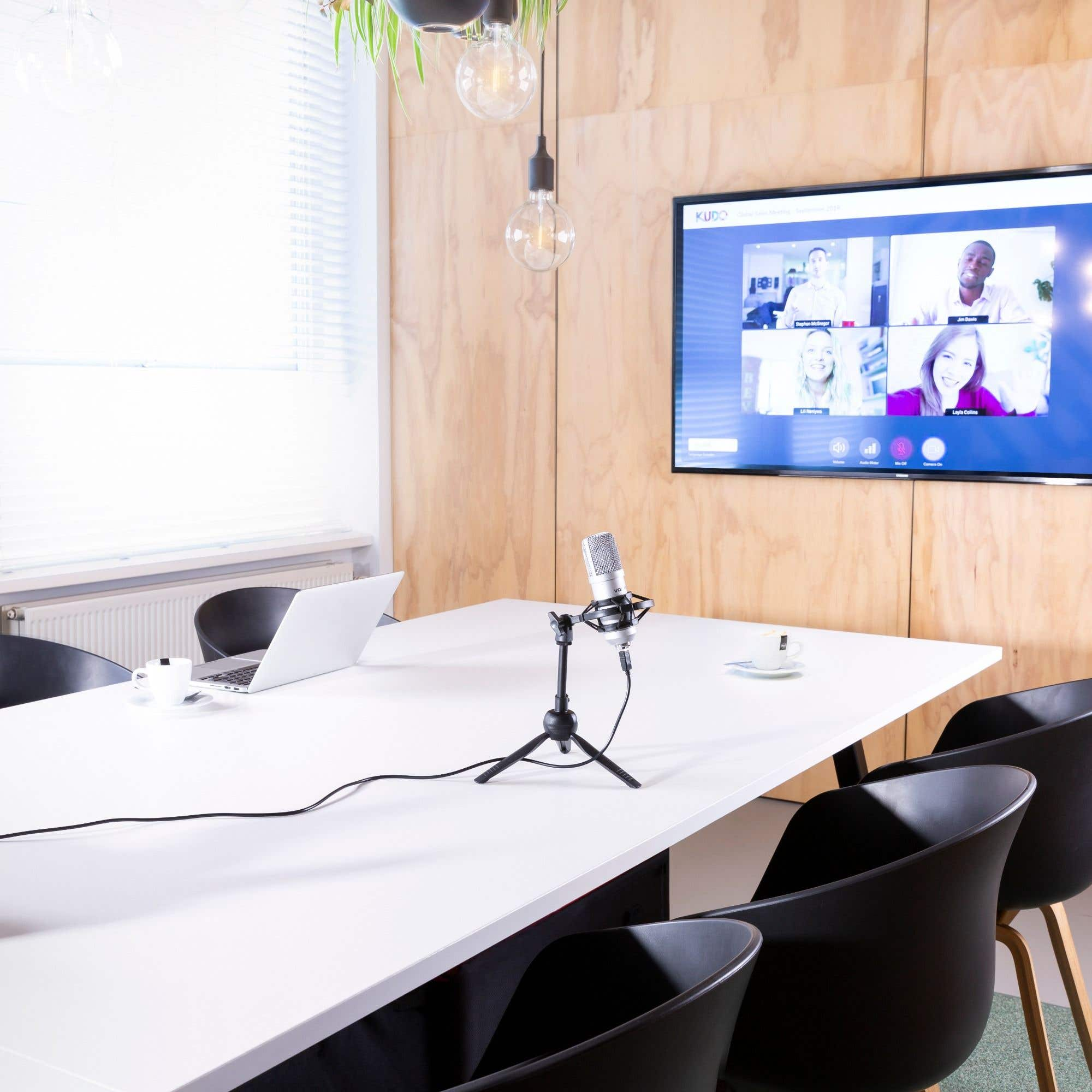 Conference room with a table, chairs, laptop, microphone, and a screen showing a video call with four people.