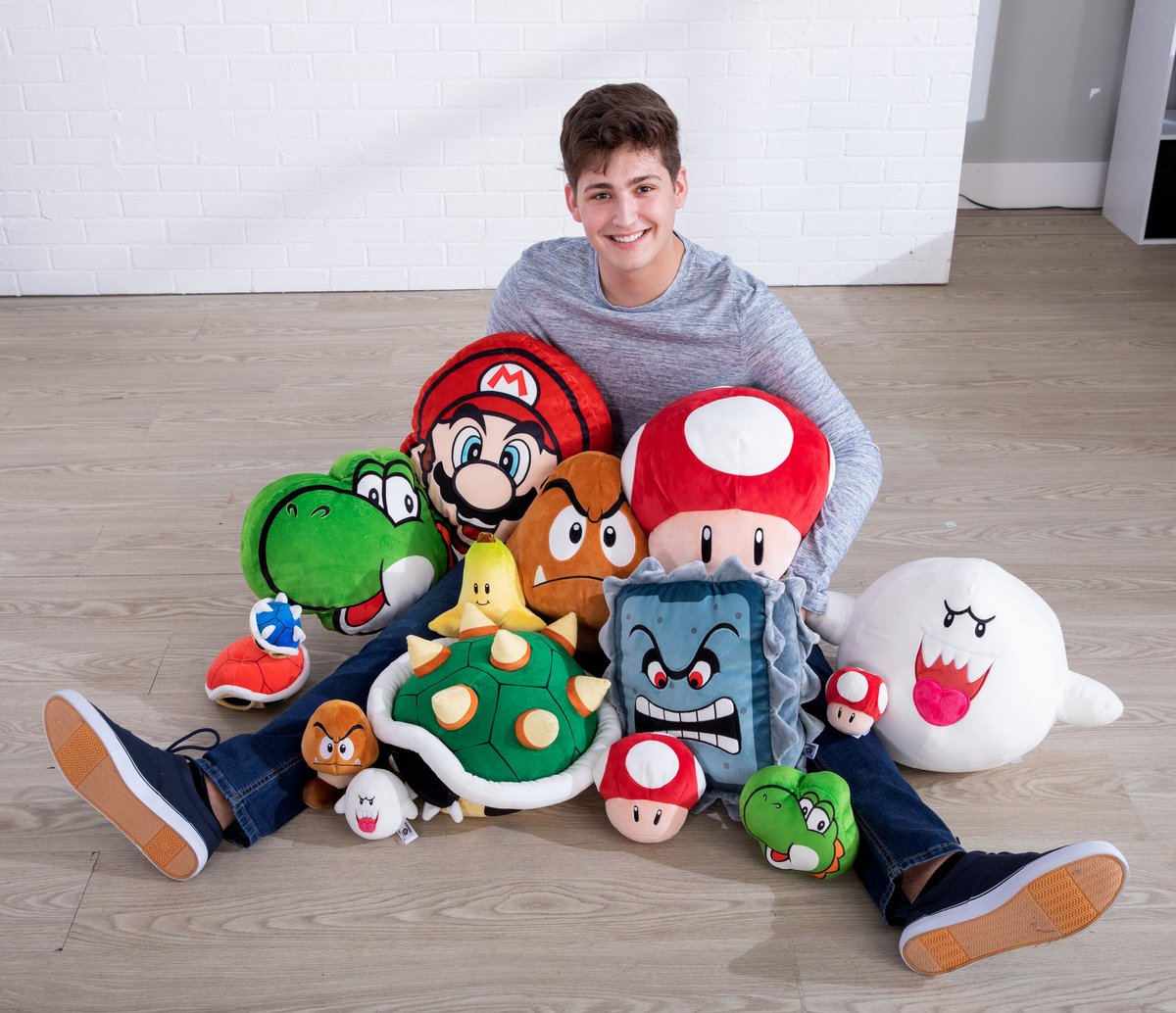 Ein lächelnder Teenager sitzt umgeben von Mario-Plüschfiguren. Ein roter Mario-Hut, Yoshi, ein Pilz und andere Charaktere sind zu sehen.