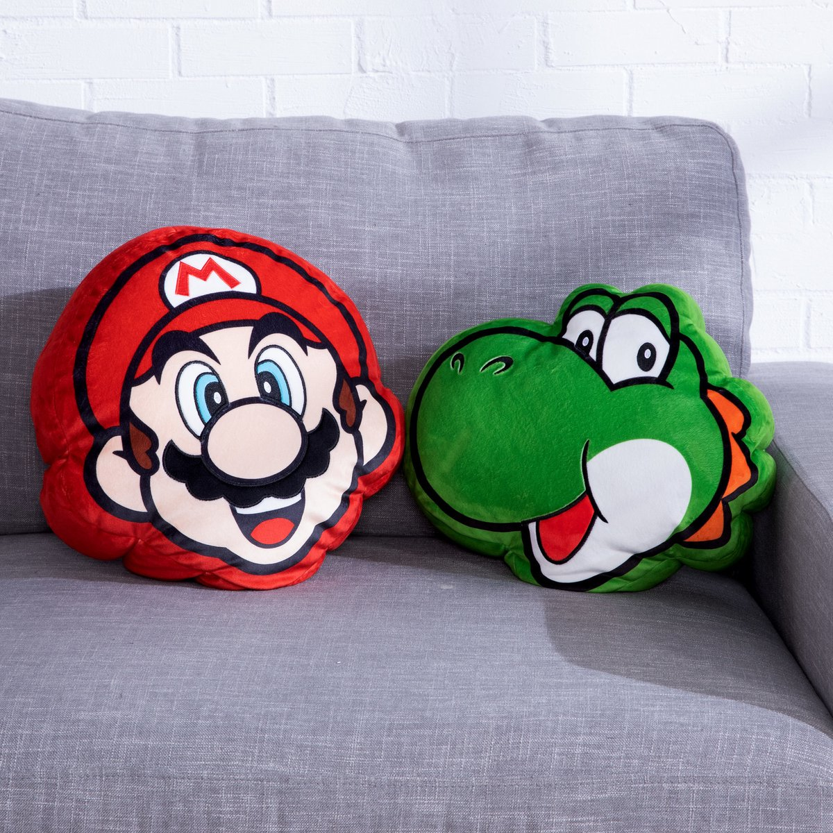 Zwei Plüschkissen: Mario mit roter Mütze und Yoshi, der grüne Dinosaurier, sitzen auf einem grauen Sofa. Im Hintergrund eine weiße Ziegelmauer.
