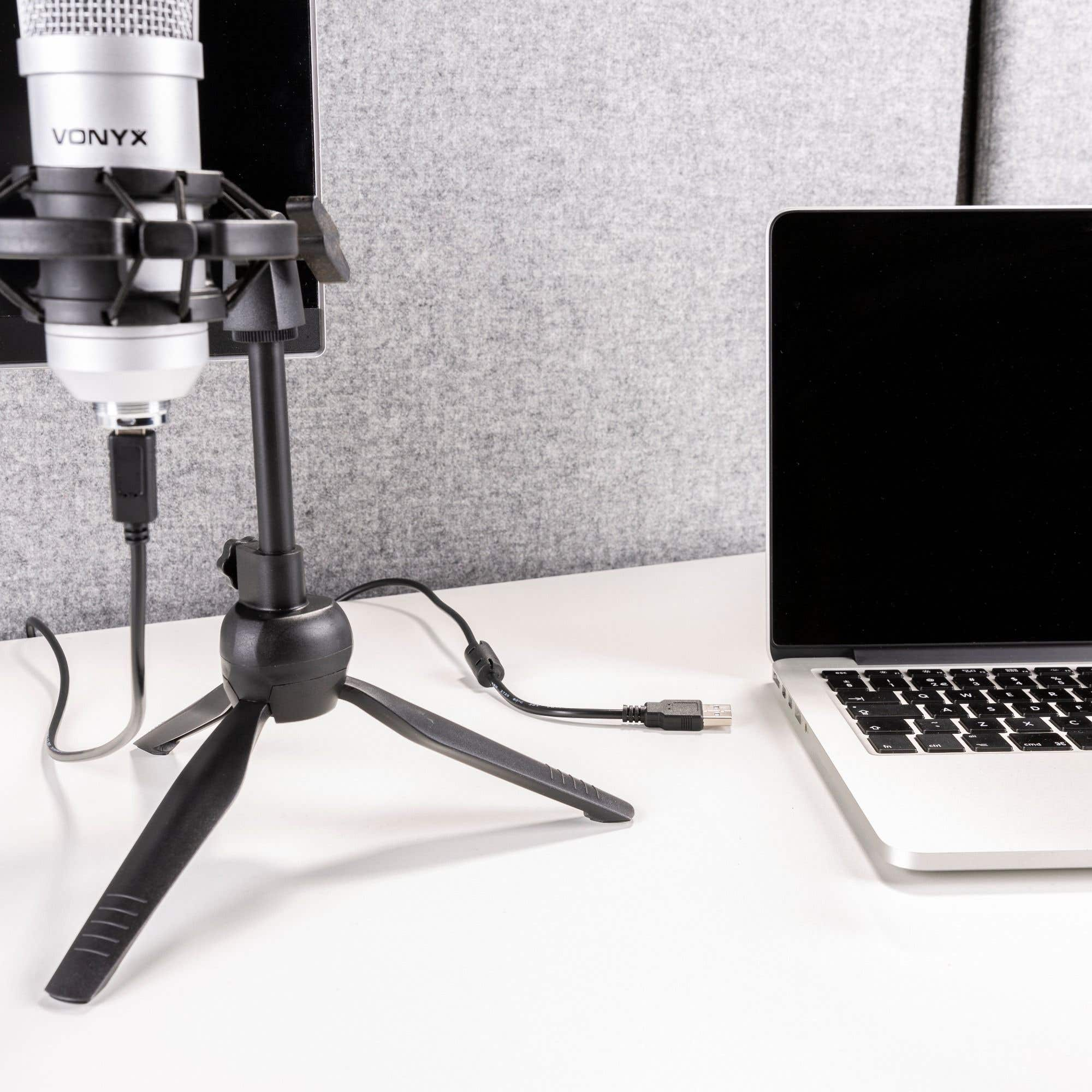 A microphone on a stand, laptop, and cables are on a white surface. Gray soundproofing in the background.