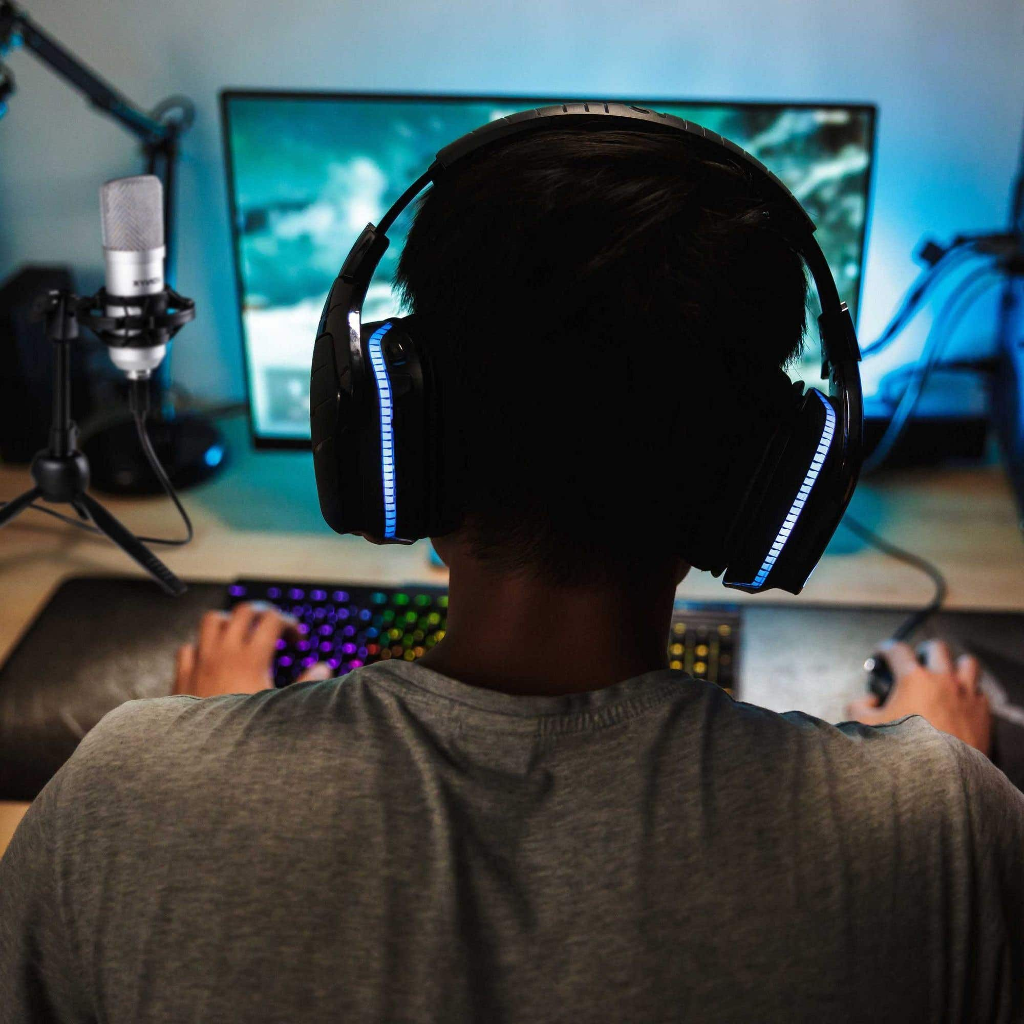 A person wearing headphones is gaming. A microphone and keyboard are in the frame. The scene is lit with blue.