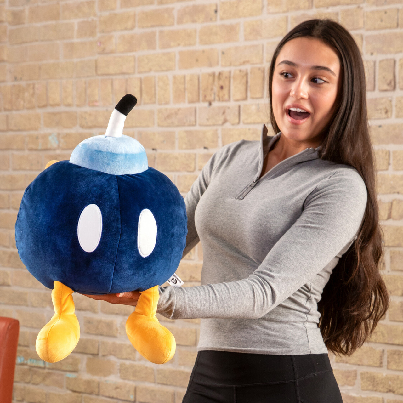 Una donna tiene in mano un peluche a forma di bomba blu con piedi gialli. Sorride sorpresa. Lo sfondo è un muro di mattoni.
