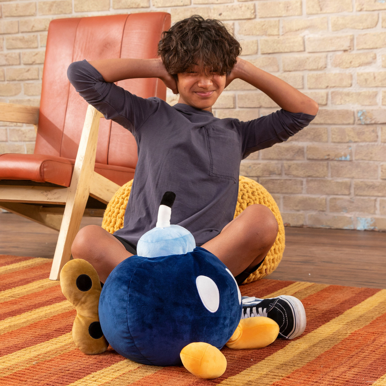 Un ragazzo seduto si copre le orecchie. Un peluche a forma di bomba blu, un peluche a forma di arachide e un cuscino giallo davanti a lui. Una sedia sullo sfondo.