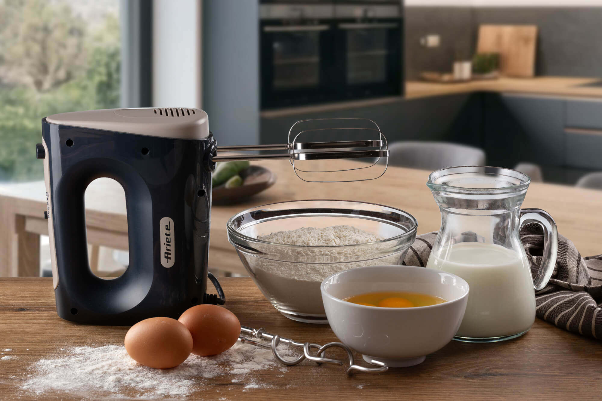 Eine Küchenszene mit Mixer, Mehl, Eiern und Milch. Der Mixer ist schwarz und cremefarben. Zutaten werden auf einem Holztisch zubereitet.