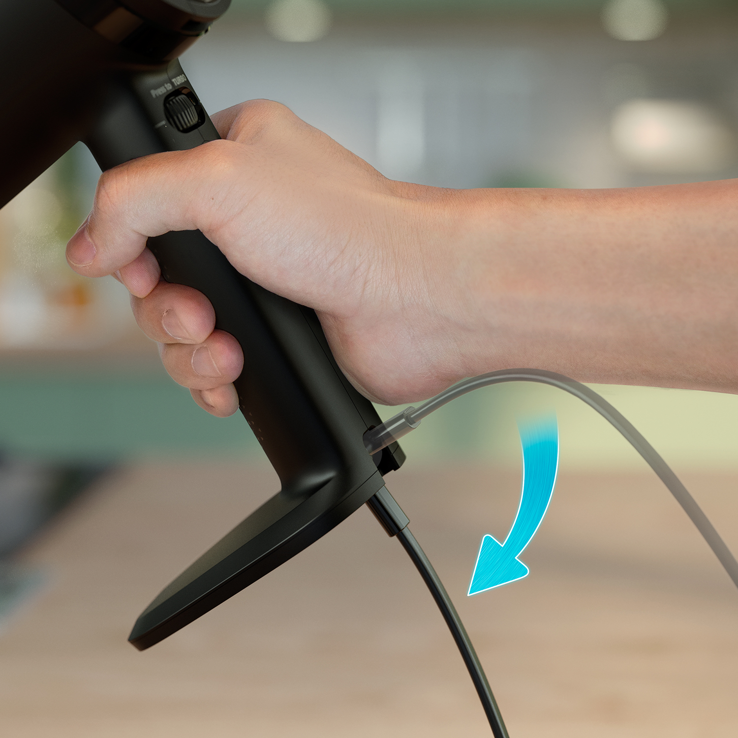Ręka trzyma czarne urządzenie z kablem. Niebieska strzałka wskazuje, że kabel jest podłączany. Tło jest rozmyte.