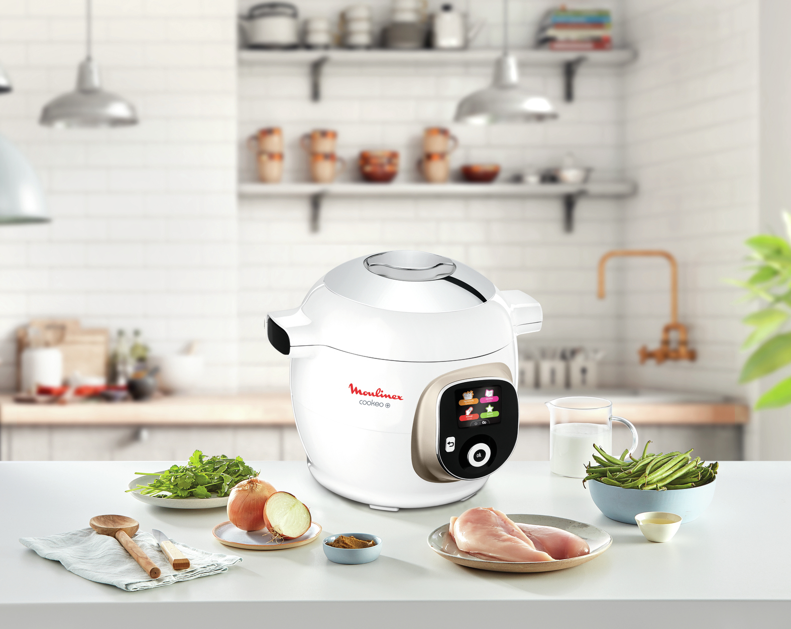 Una olla a presión Moulinex Cookeo blanca está sobre una mesa con ingredientes. Incluye pollo, verduras y un vaso de leche.