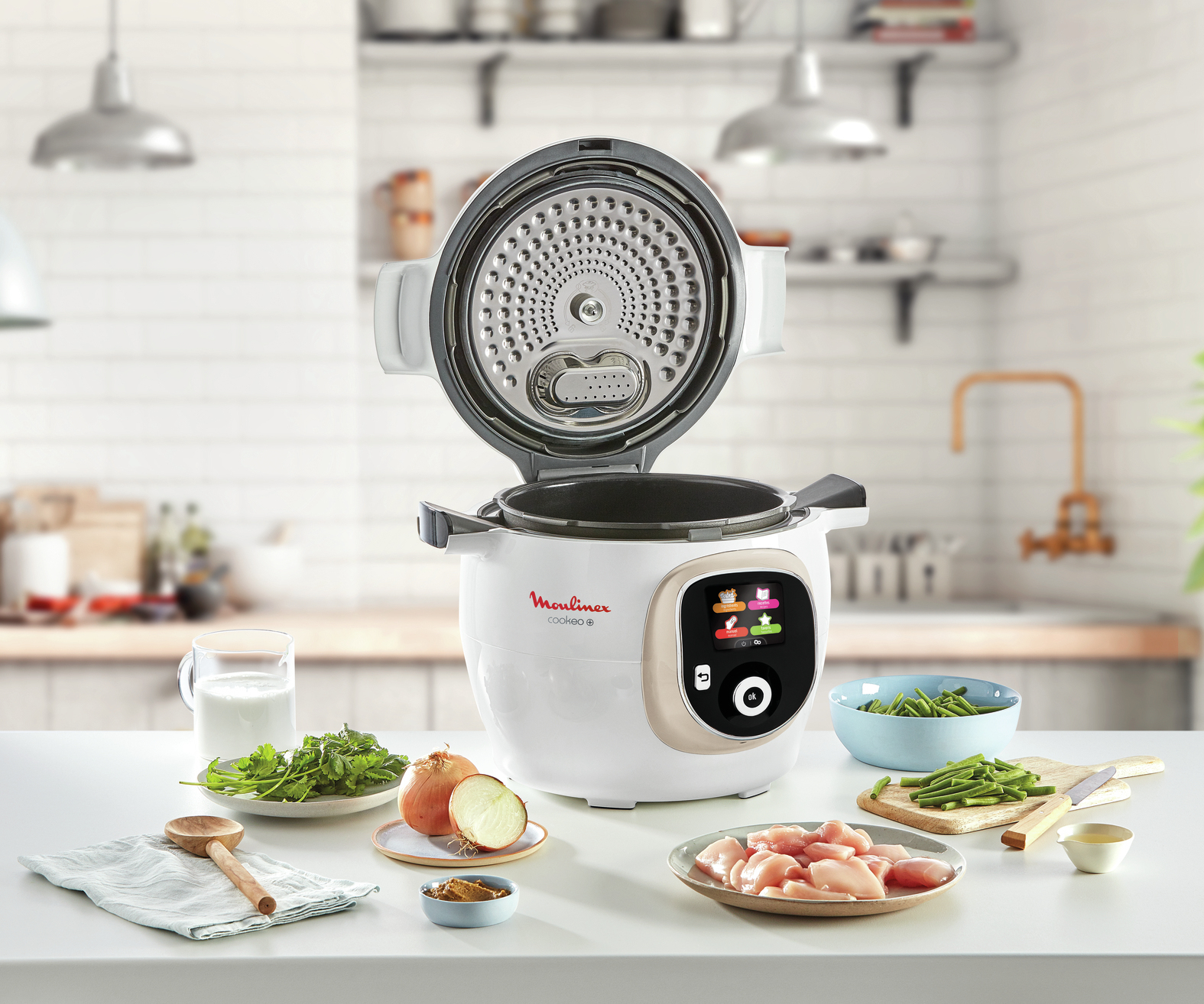 Un Moulinex Cookeo blanco sobre una mesa con ingredientes: pollo, cebollas, judías verdes, hierbas y leche. La tapa de la olla está abierta.