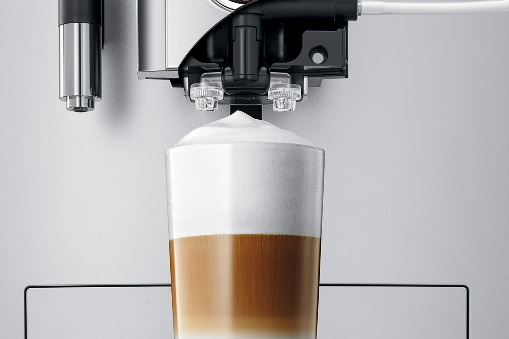 Nahaufnahme eines Glases geschichteten Latte Macchiato mit Milchschaum. Kaffeemaschinenteile befinden sich über dem Glas, im Hintergrund eine graue Wand.