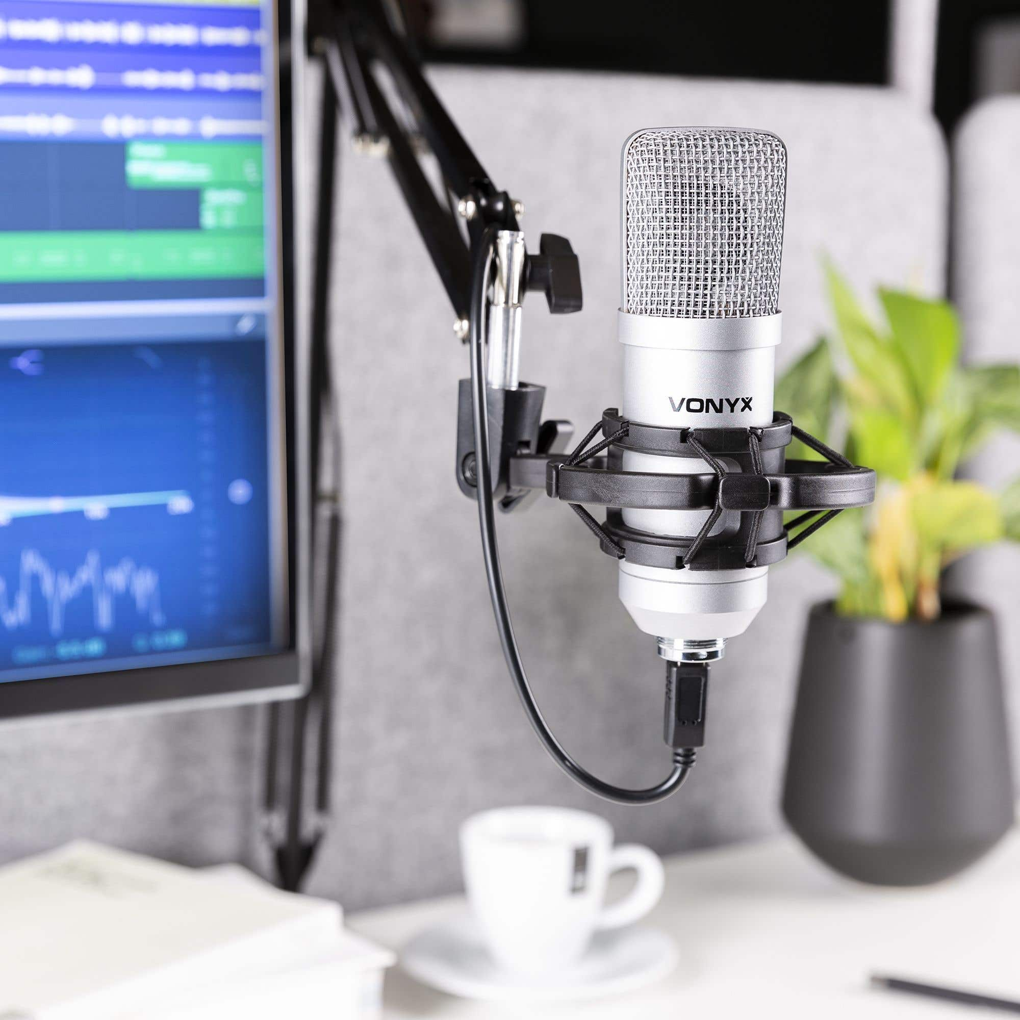 A silver microphone with a black stand is in front of a computer screen and a plant. A coffee cup is in the foreground.