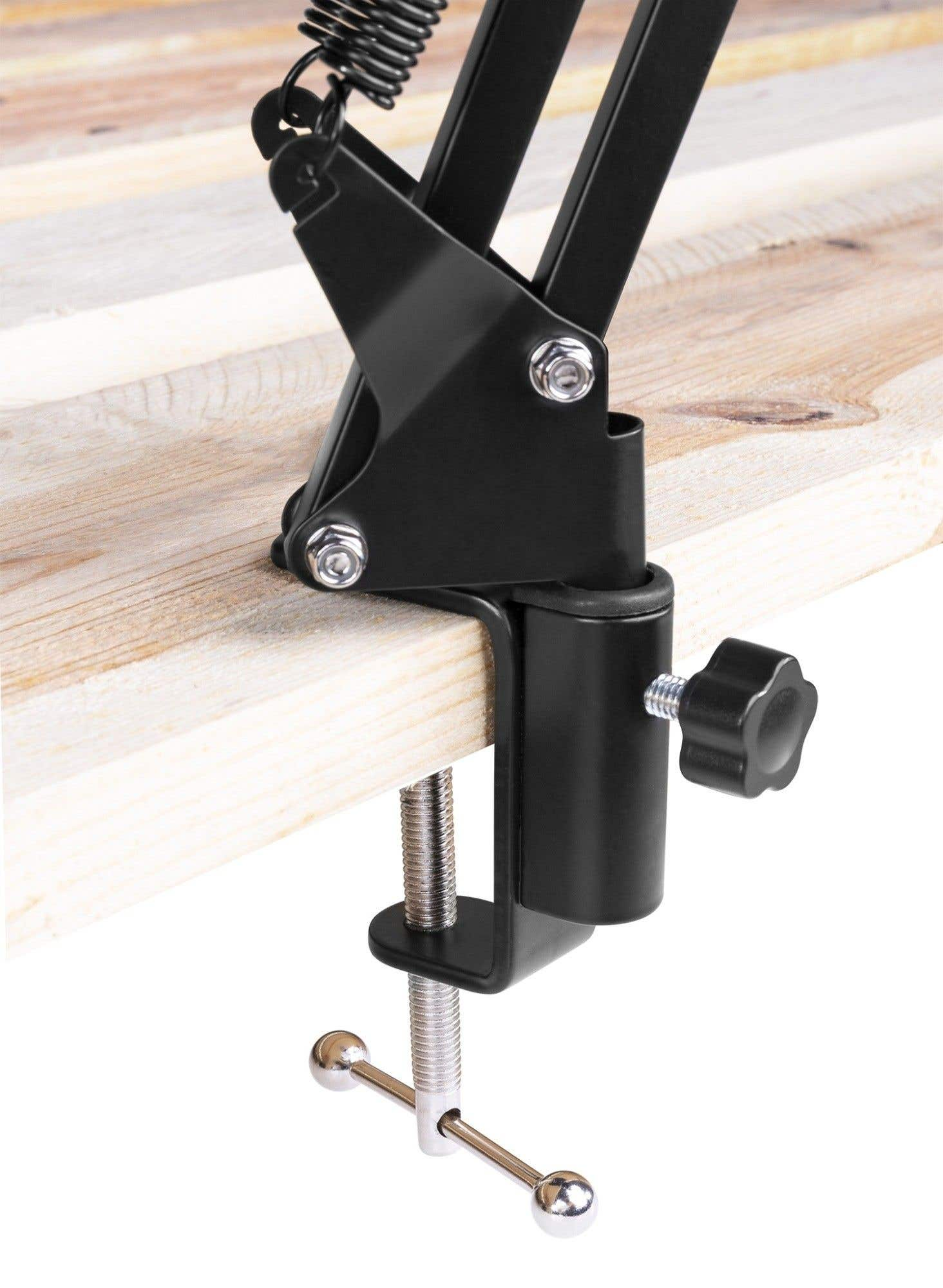 A black metal clamp, attached to a wooden surface. A spring and arm are visible. The clamp includes a screw and a ball joint.