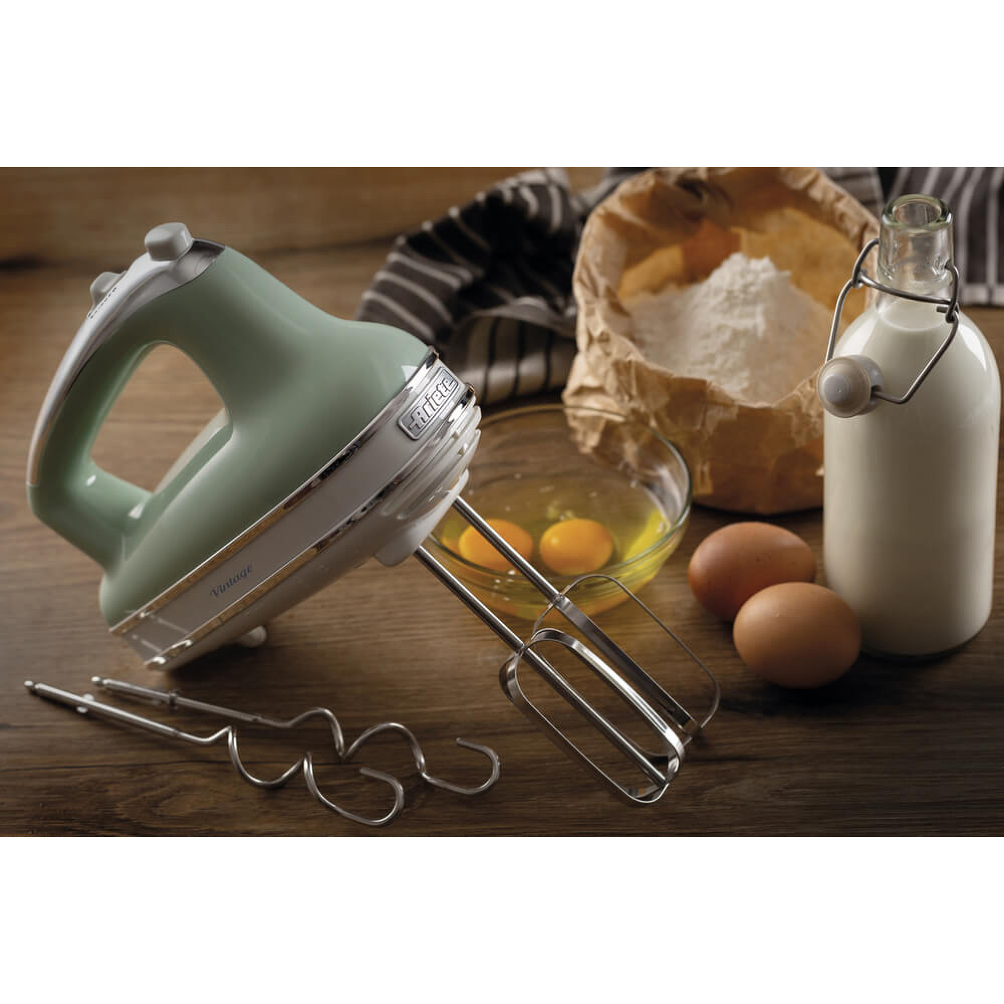 Grüner und weißer Handmixer, Eier, Mehl, Milchflasche auf Holzoberfläche. Backzutaten sind angeordnet.