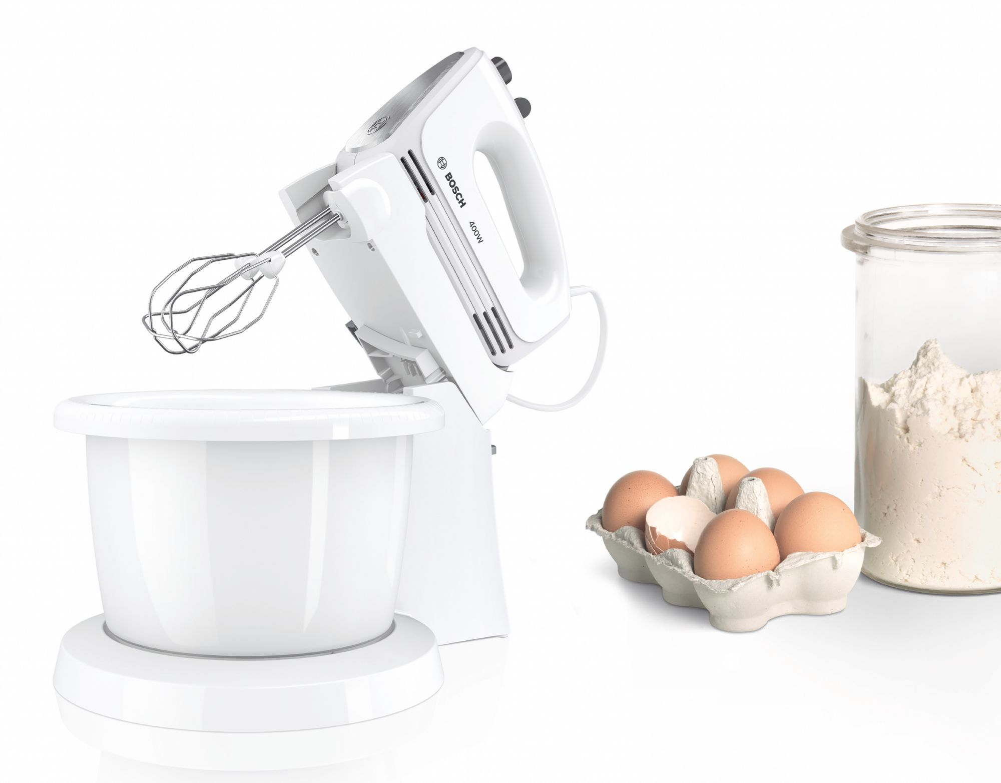 Ein weißer Bosch-Handmixer mit Schüssel, Eiern im Karton und Mehl in einem Glas auf weißer Oberfläche. Backzutaten.