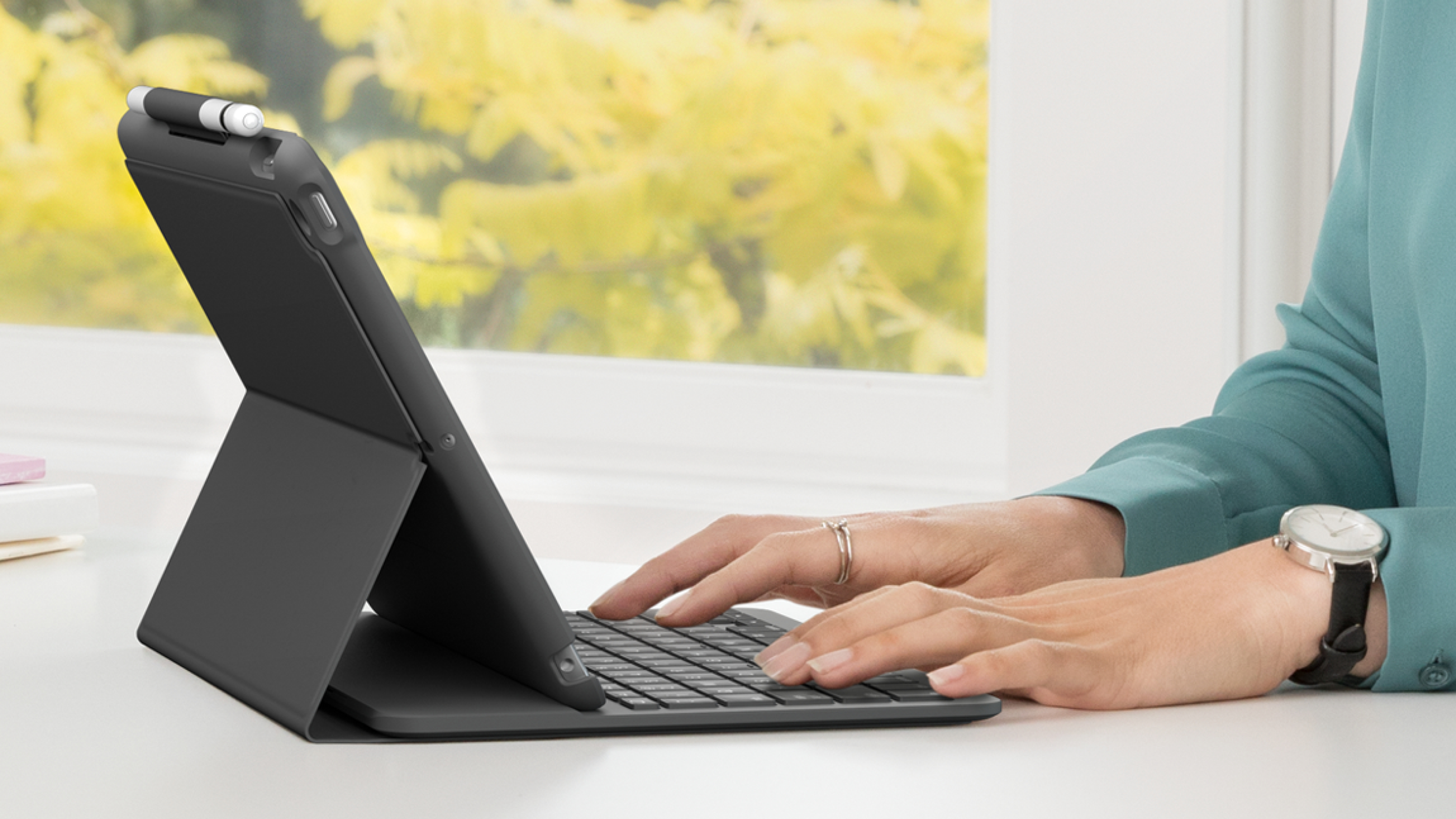 Una persona digita su una tastiera nera per tablet. Una penna è appoggiata sul tablet. Lo sfondo è una finestra con fogliame sfocato.