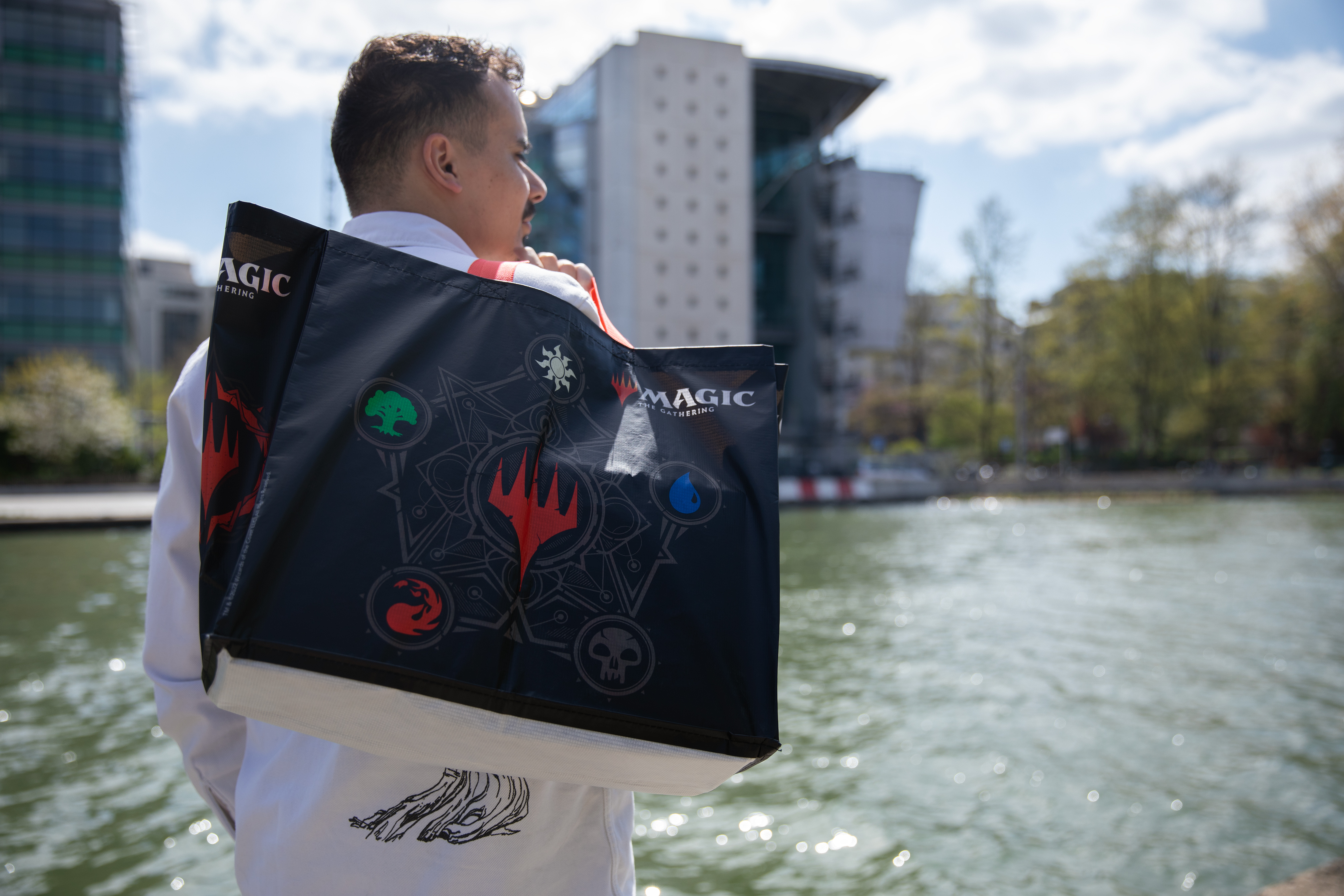 Hombre con bolsa de Magic, de pie cerca del agua. Edificios y árboles al fondo.