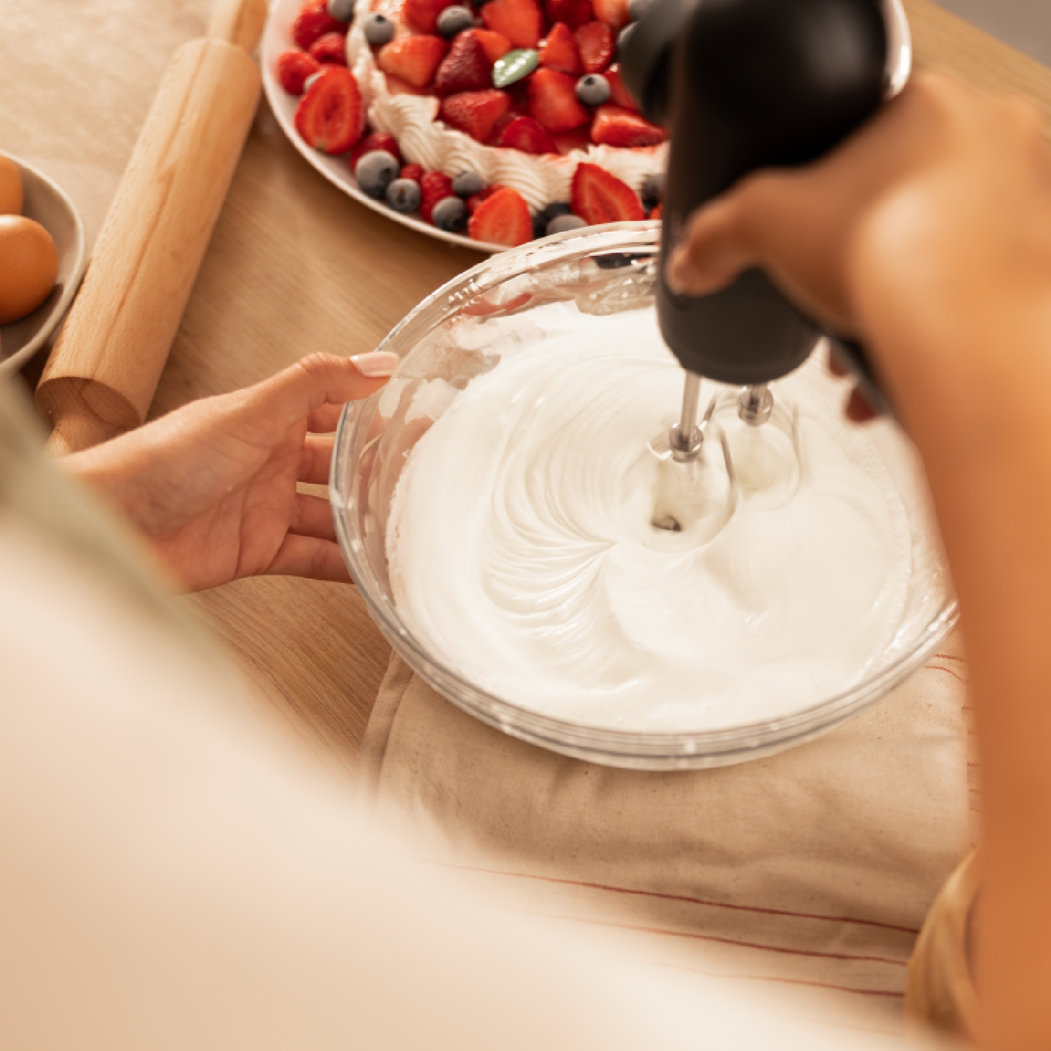 Una persona sta mescolando la panna in una ciotola di vetro con un frullatore elettrico, torta con frutti di bosco sullo sfondo.