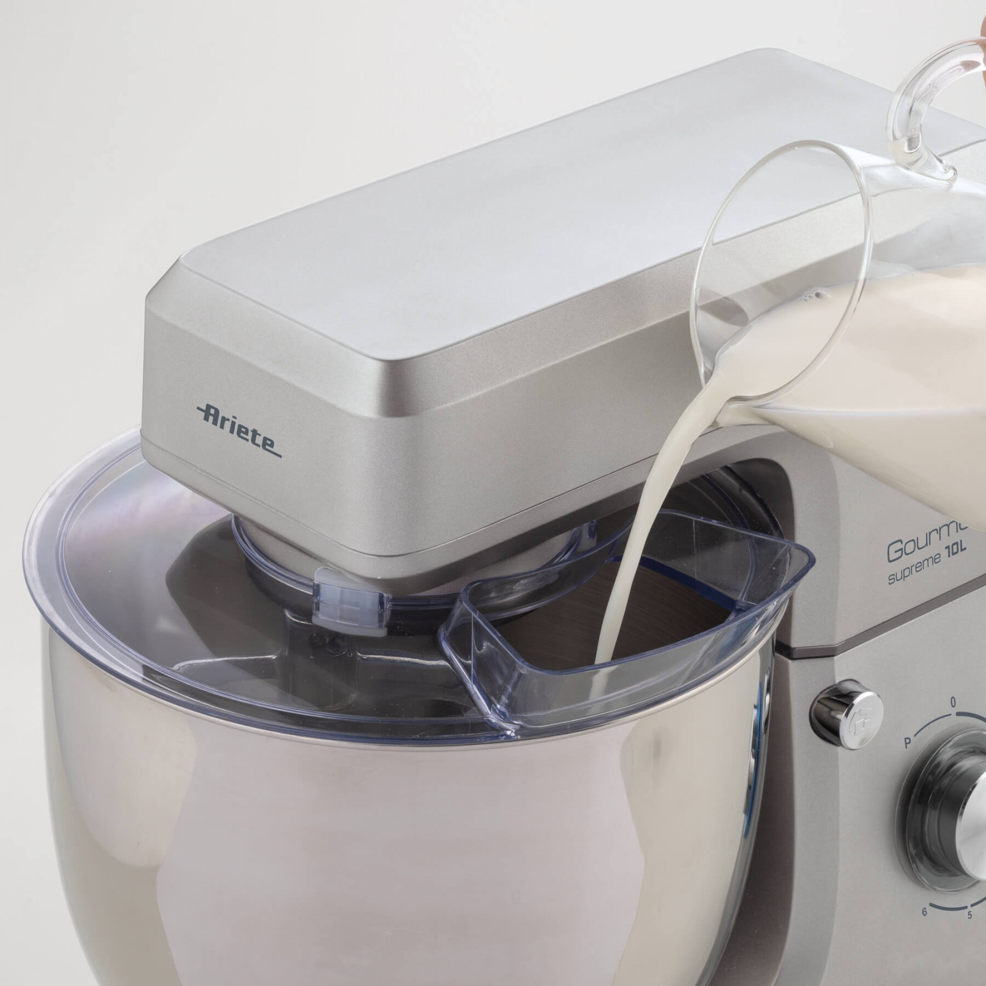 Un'impastatrice Ariete argentata viene riempita di latte da una brocca di vetro. Coperchio in plastica trasparente. Sfondo bianco.