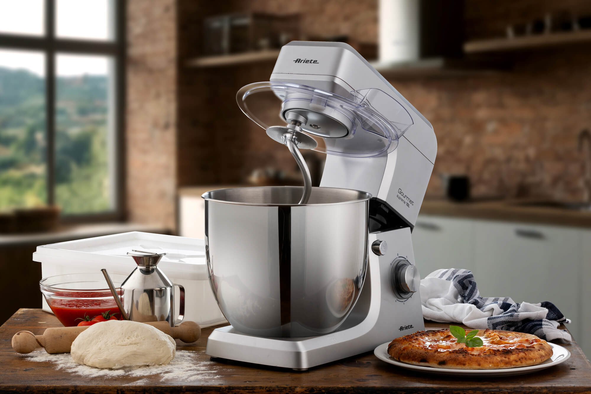 Scena di cucina con impastatrice, impasto, pizza e ingredienti su un tavolo di legno. Sfondo con finestra e armadi.