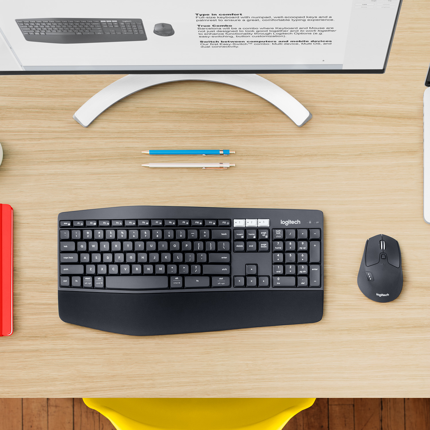 Biurko z klawiaturą, myszą, monitorem i długopisami. Klawiatura i mysz są czarne. Biurko jest w kolorze drewna.