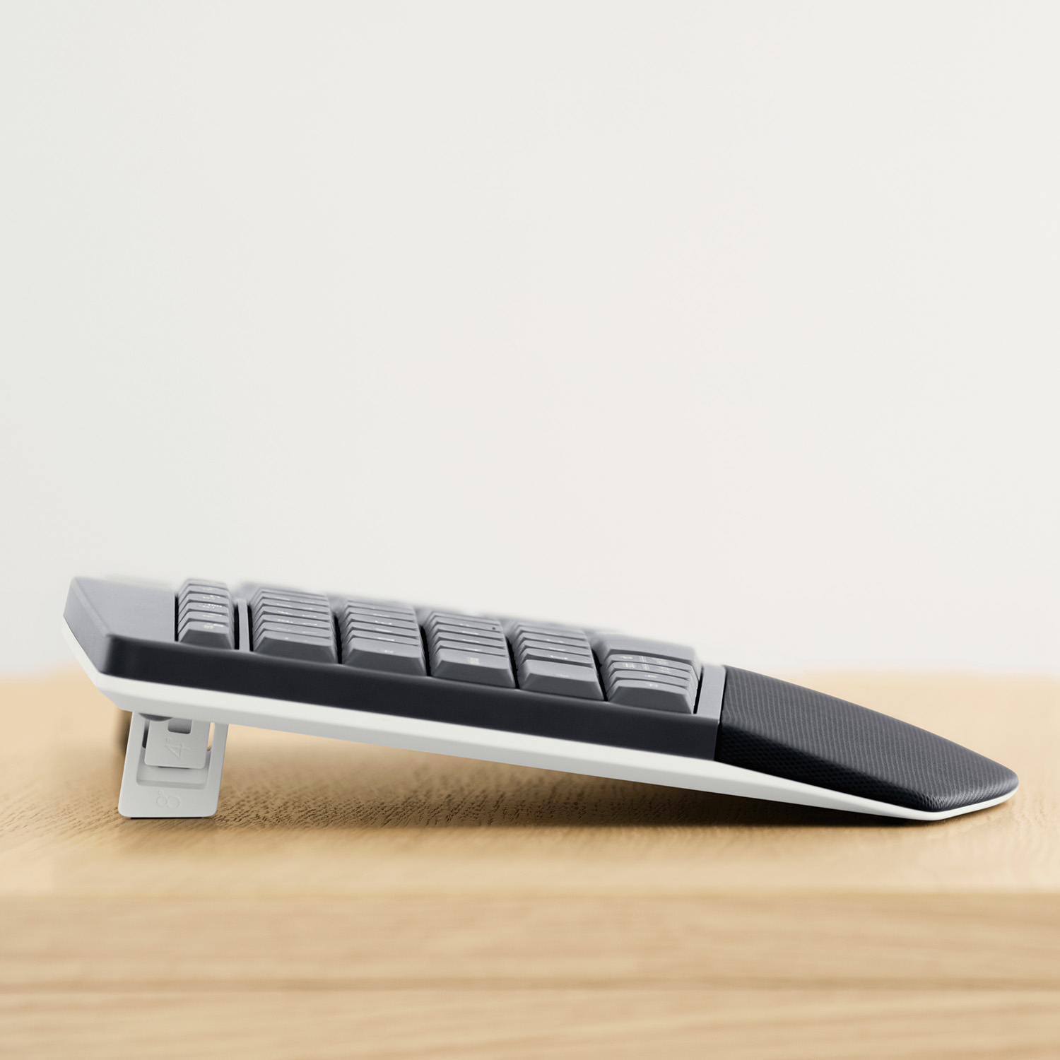 Widok z boku podzielonej ergonomicznej klawiatury na drewnianej powierzchni. Klawiatura jest szara i biała, z czarną podpórką pod nadgarstek.