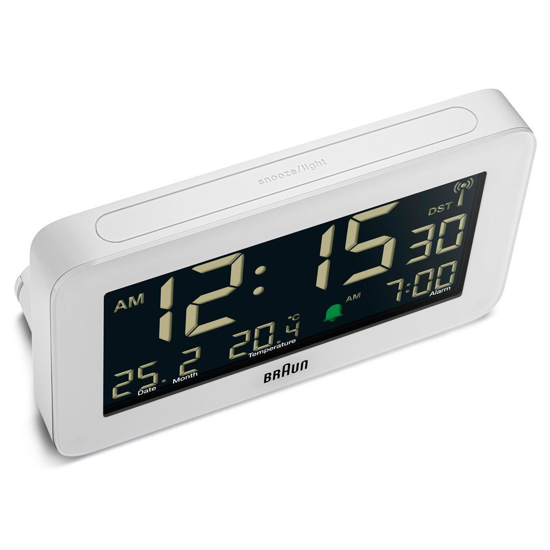 Weißer digitaler Braun-Wecker mit Anzeige von 12:15:30 Uhr morgens, Datum, Temperatur und Weckzeit 7:00. Die Schlummer-/Licht-Taste ist sichtbar.