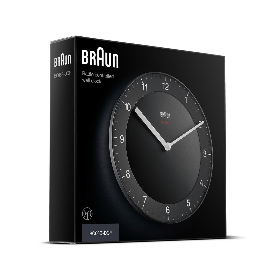 Eine schwarze, funkgesteuerte Wanduhr von Braun in ihrem Karton. Das Zifferblatt ist schwarz mit weißen Zeigern und Zahlen. Die Box ist schwarz.