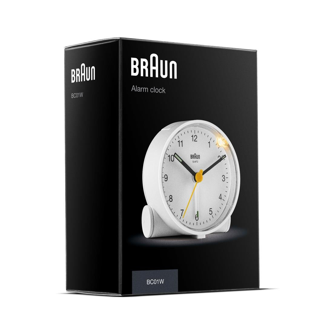 Ein weißer Braun-Wecker in einer schwarzen Schachtel. Die Uhr hat schwarze Zeiger und einen gelben Sekundenzeiger. Die Zeit ist ca. 10:02.