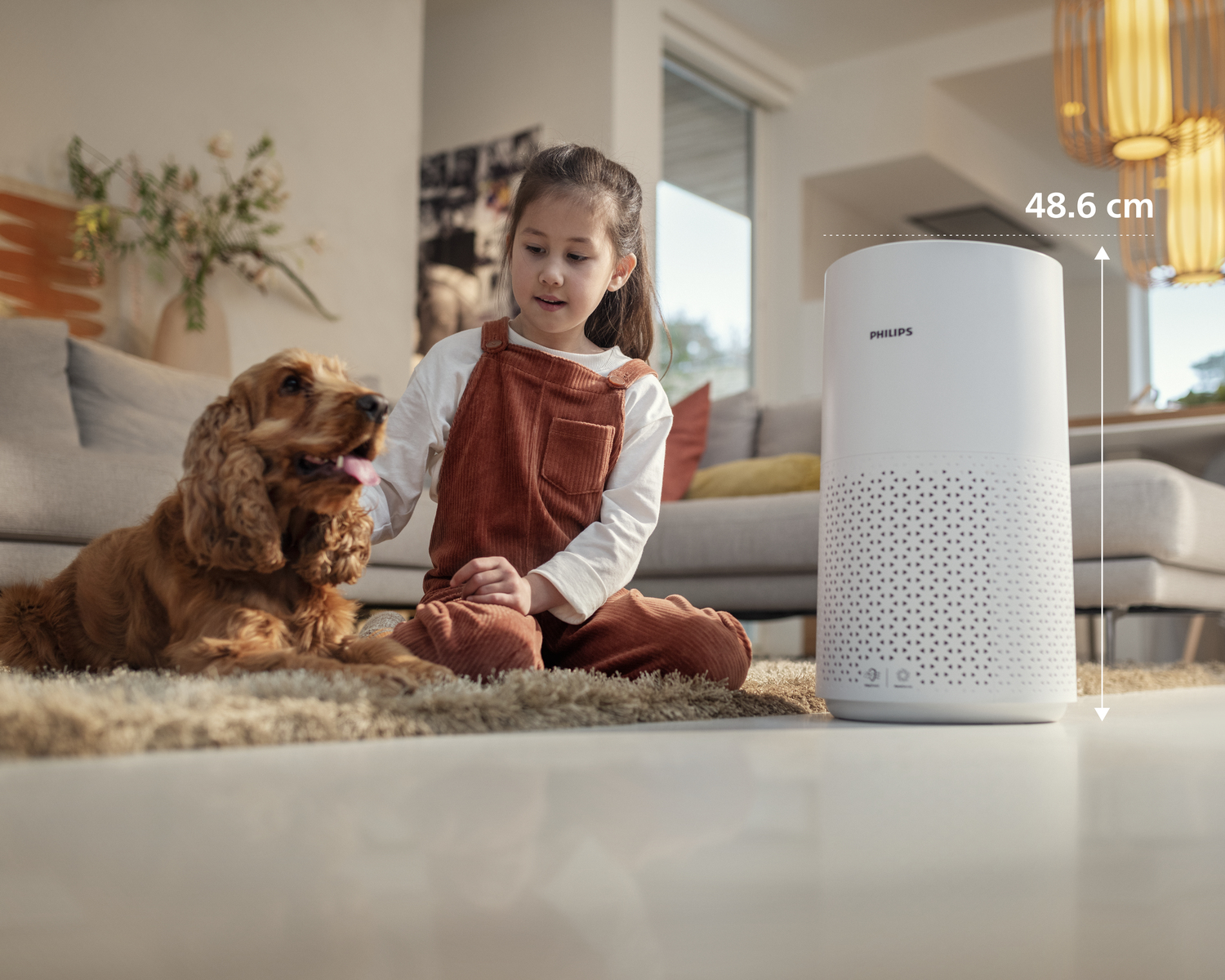Bir kız ve bir köpek, beyaz bir Philips hava temizleyicinin yanında yerde oturuyor. Köpek kahverengi ve kız tulum giyiyor.