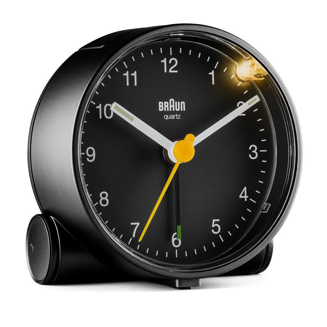 Un réveil Braun à quartz noir. Il a un cadran noir avec des chiffres et des aiguilles blanches. Une trotteuse jaune. Une lumière est allumée.