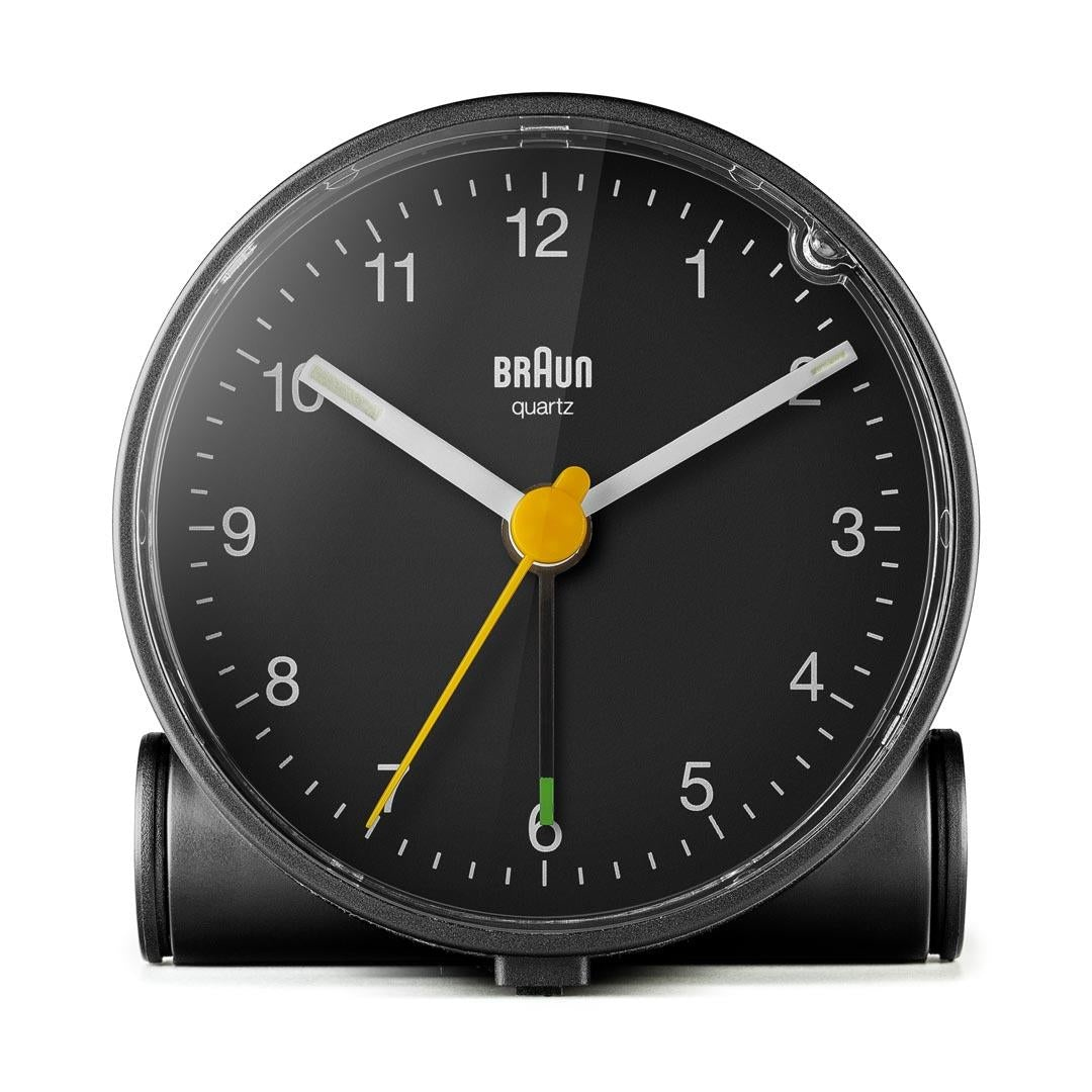 Un réveil Braun quartz noir. Il a un cadran noir avec des aiguilles et des marqueurs blancs. L'aiguille des secondes est jaune. L'horloge est sur fond blanc.