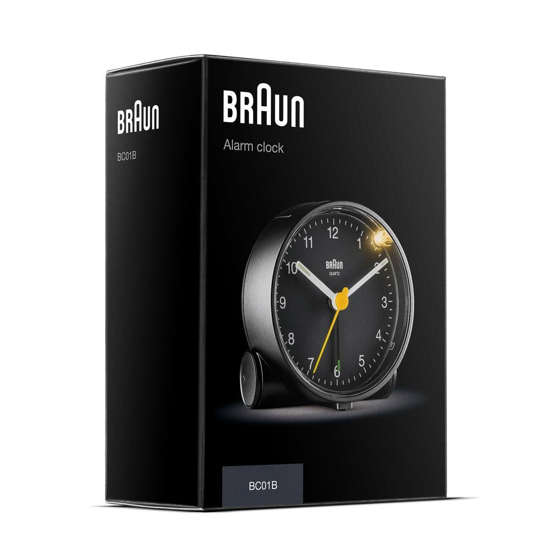 Un réveil Braun dans une boîte noire. Le cadran est noir avec des chiffres blancs et des aiguilles jaunes. La boîte est noire.