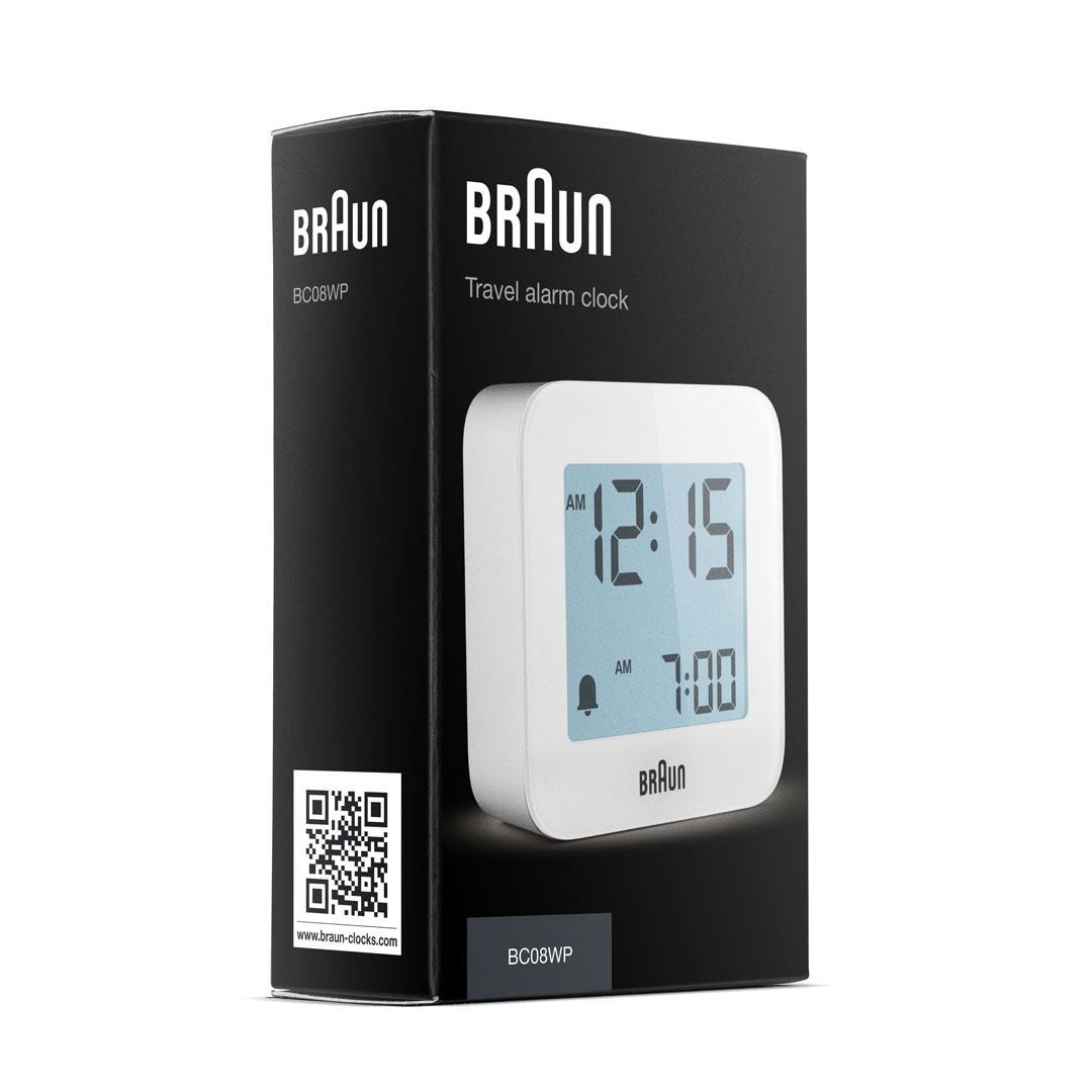 Ein Braun Reise-Wecker in seiner Box. Die Uhr zeigt 12:15 Uhr und 7:00 Uhr an. Die Box hat das Braun-Logo und das Modell BC08WP.