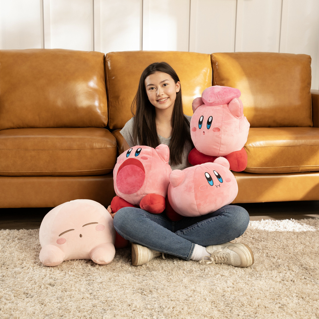 Ein Mädchen sitzt, umgeben von rosa Kirby-Plüschtieren. Hinter ihr steht ein braunes Sofa. Der Boden ist mit einem beigen Teppich bedeckt.