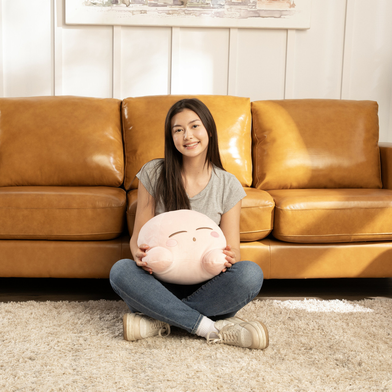 Eine junge Frau sitzt auf einem Teppich und hält ein rosa Plüschtier. Hinter ihr ist ein braunes Ledersofa. Sie trägt ein graues Hemd und Jeans.