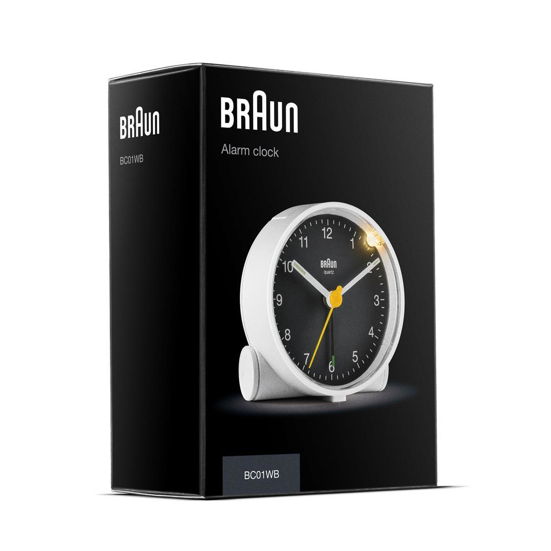 Ein weißer Braun-Wecker in seiner schwarzen Schachtel. Die Uhr hat ein schwarzes Zifferblatt mit weißen Zahlen und gelben Zeigern.