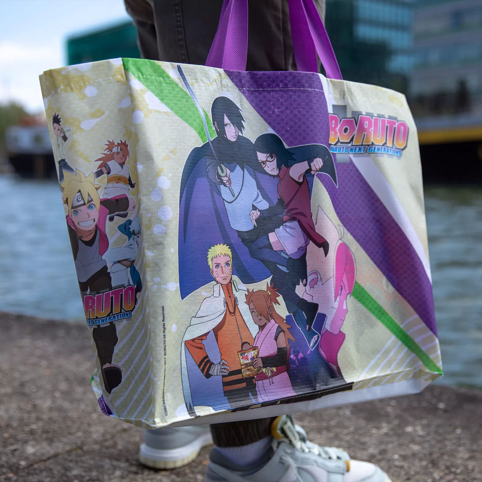 Eine Person hält eine Tragetasche mit Boruto-Charakteren. Der Hintergrund umfasst Wasser und ein Gebäude.